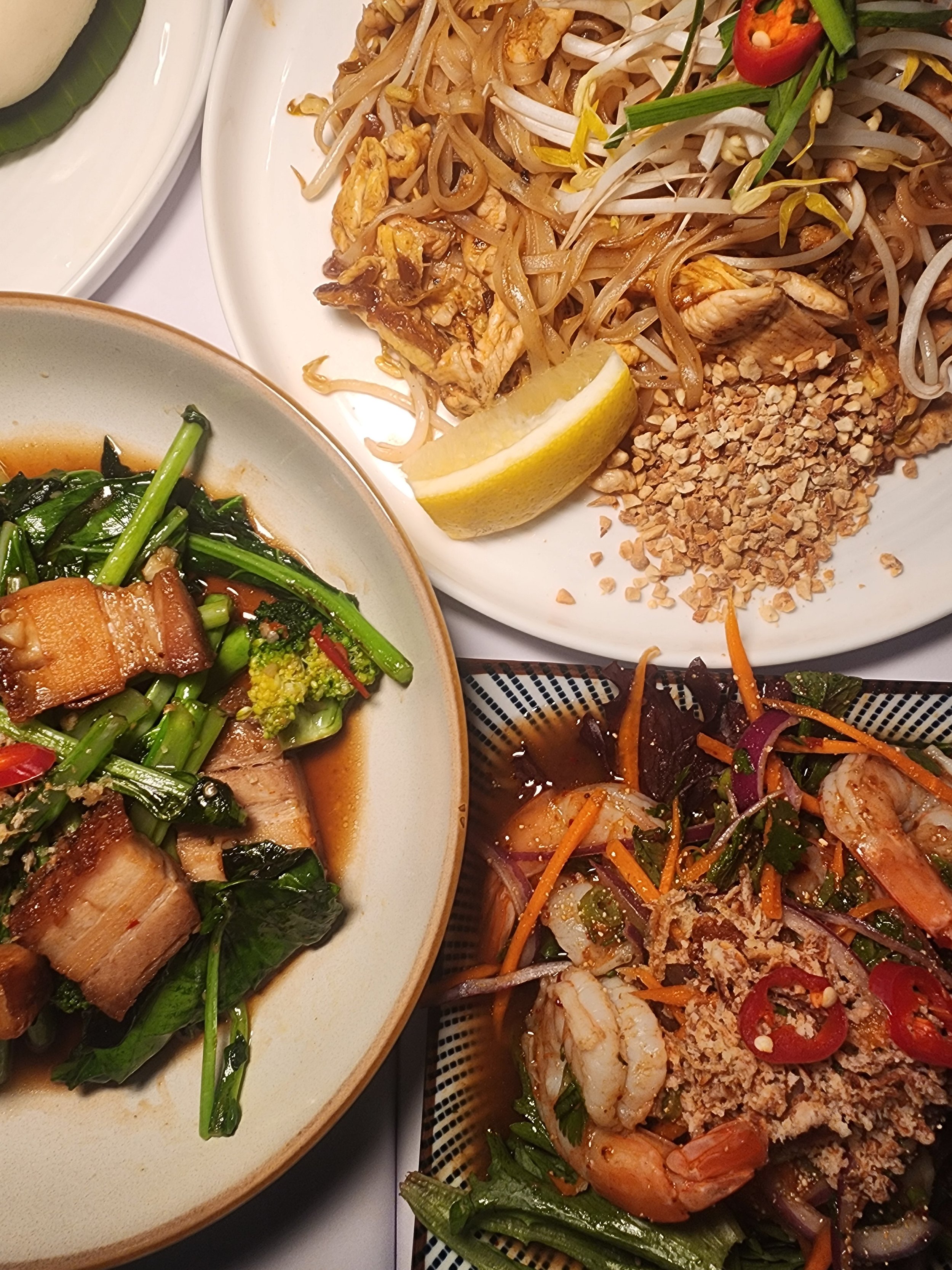 Three plates of Asian cuisine including noodles with chicken and peanuts, stir-fried vegetables with bacon, and a shrimp salad with vegetables and chili slices.