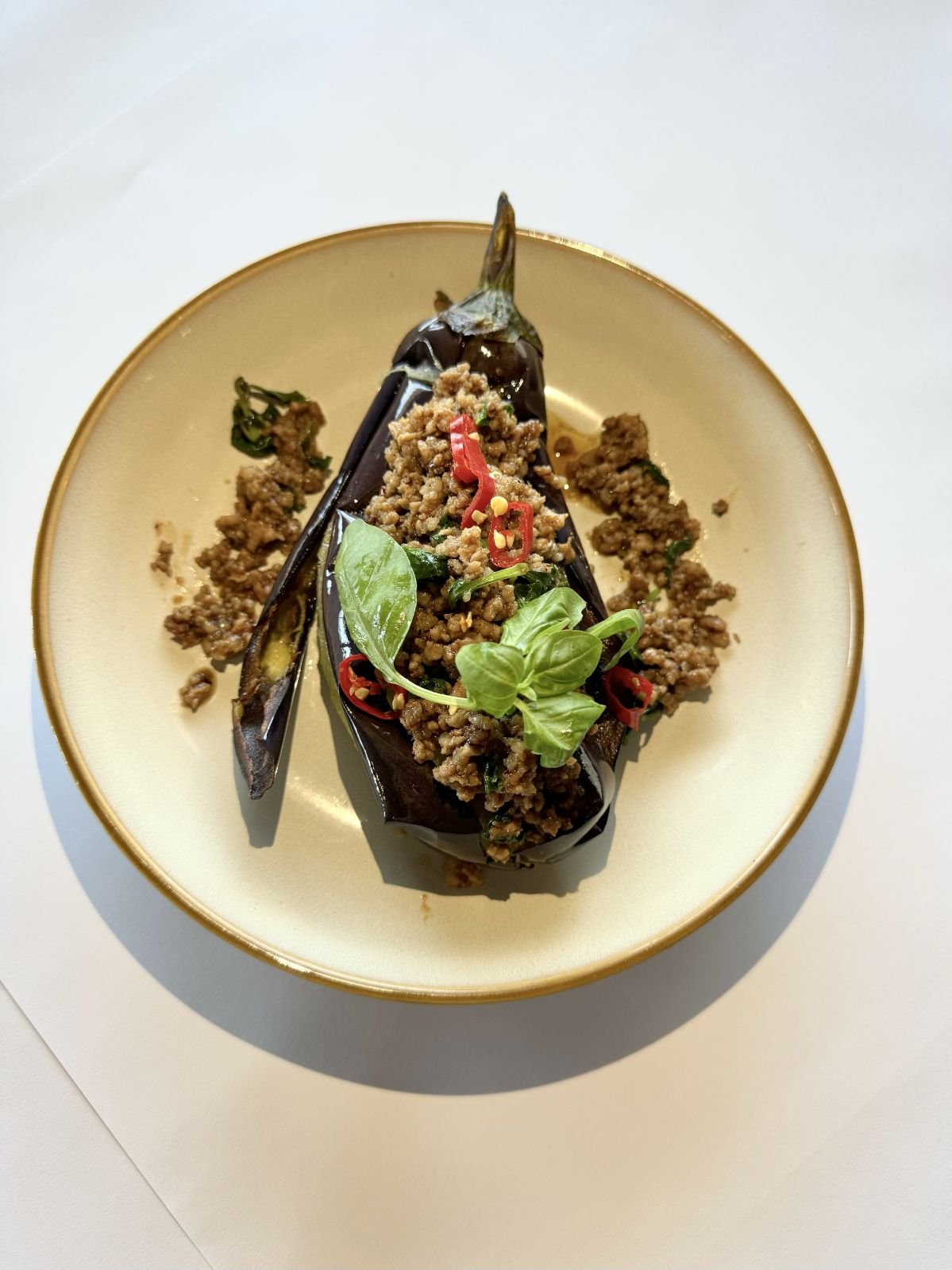 Stuffed eggplant topped with Kapow, chopped red chili, and basil leaves, served on a white plate.