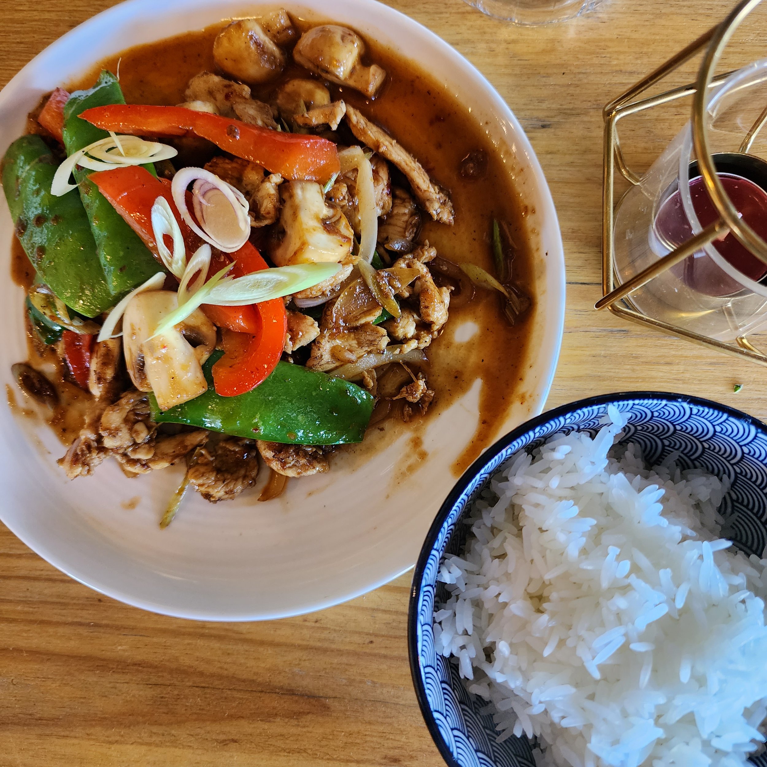 A meal consisting of a bowl of stir-fried vegetables and meat with sauce, a bowl of white rice, and a small glass of red wine on a wooden table.