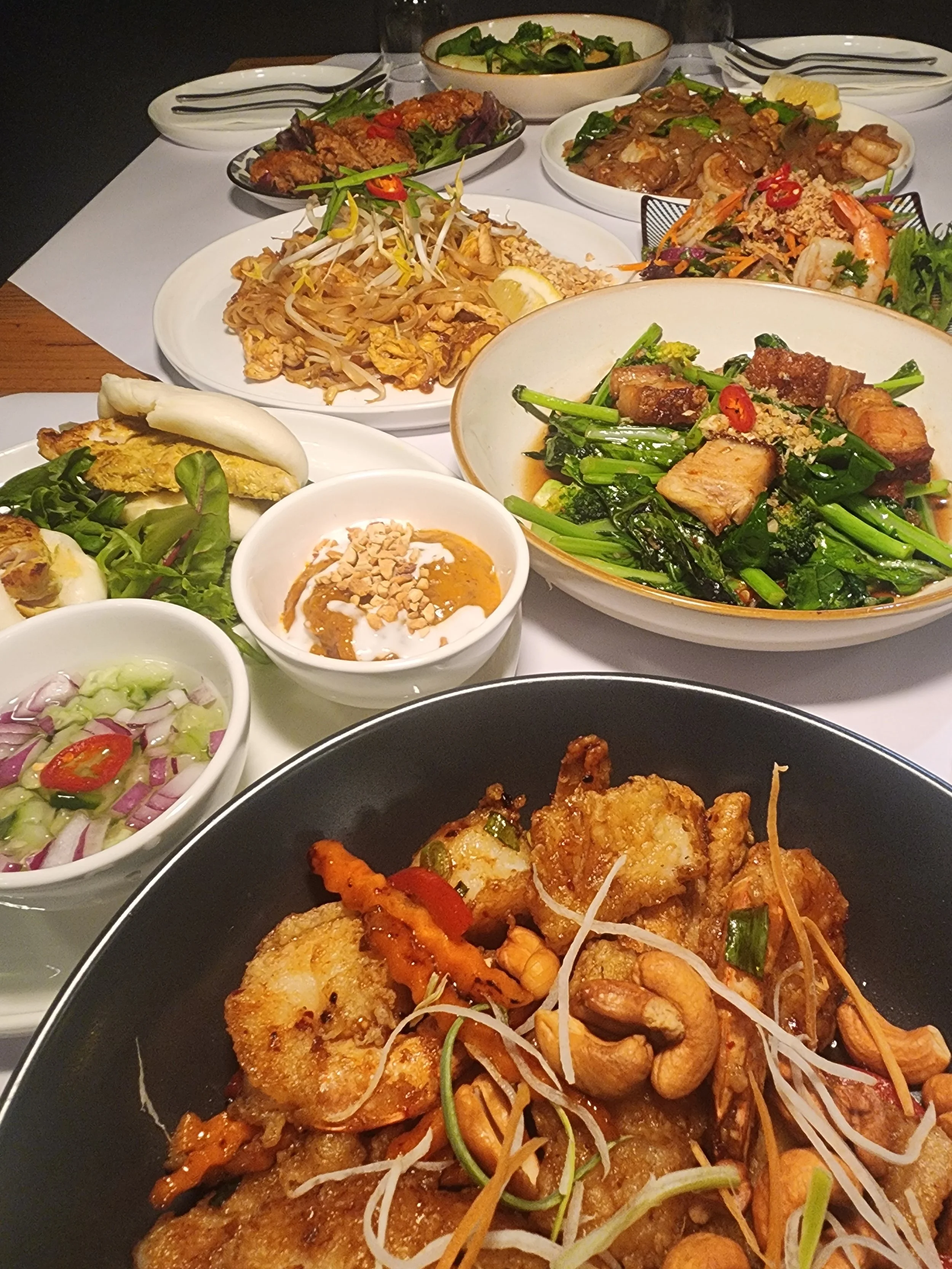 Assorted Asian dishes on a table, including Pad Thai or fish with cashews, stir-fried vegetables, buffalo chicken wings, and various dipping sauces.