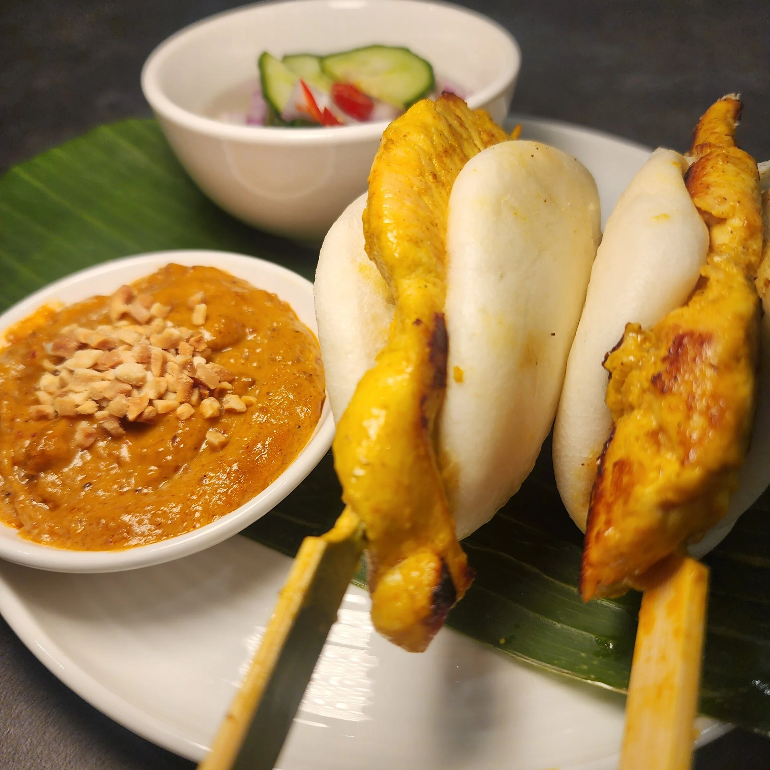 Close-up of two chicken skewers with Roti bread, served with peanut sauce and a side salad of cucumber, tomato, and onion.