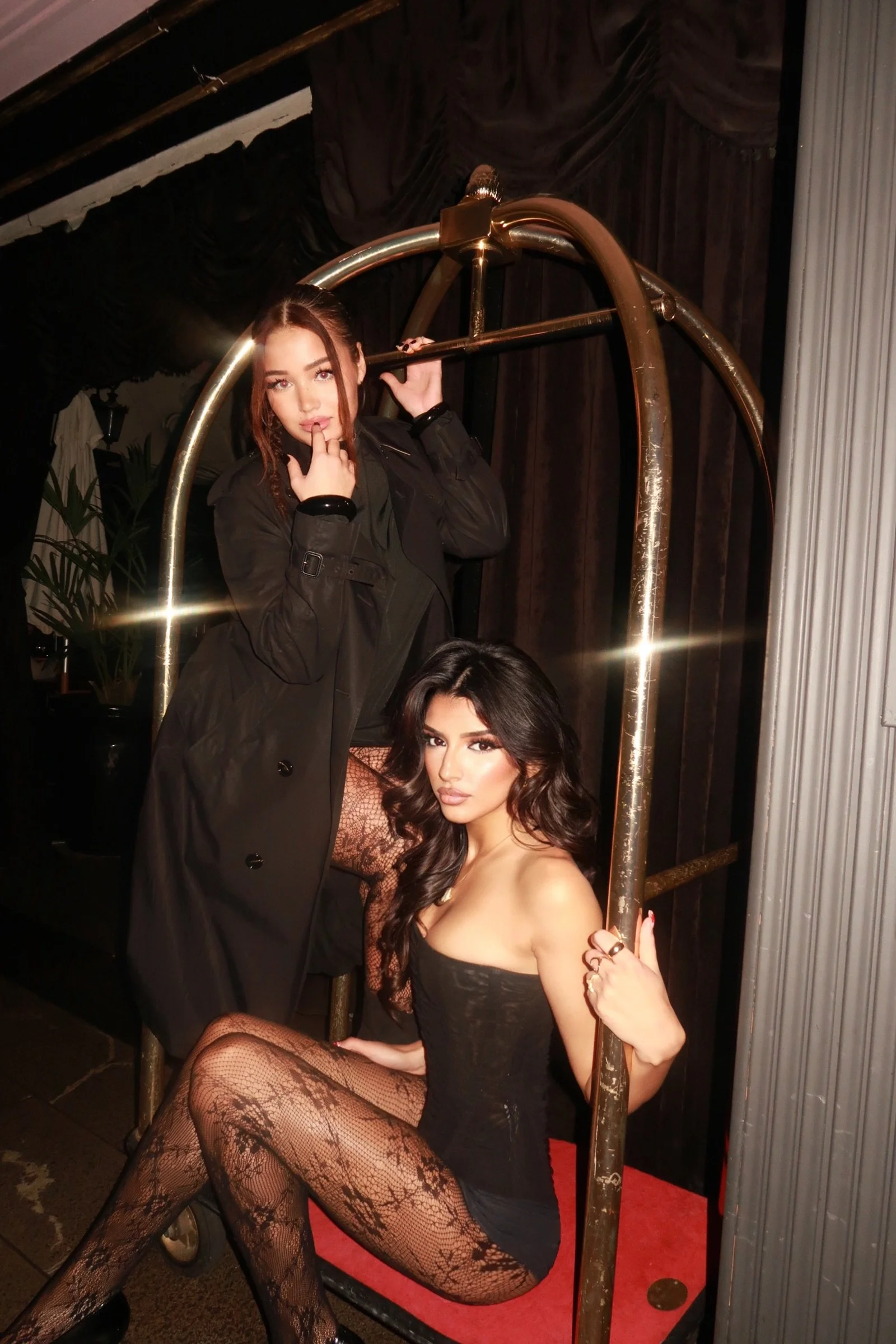 Two women posing on a vintage-style luggage cart, one standing and the other sitting, both dressed in fashionable dark clothing with fishnet stockings, in a dimly lit room with dark curtains and plants in the background.