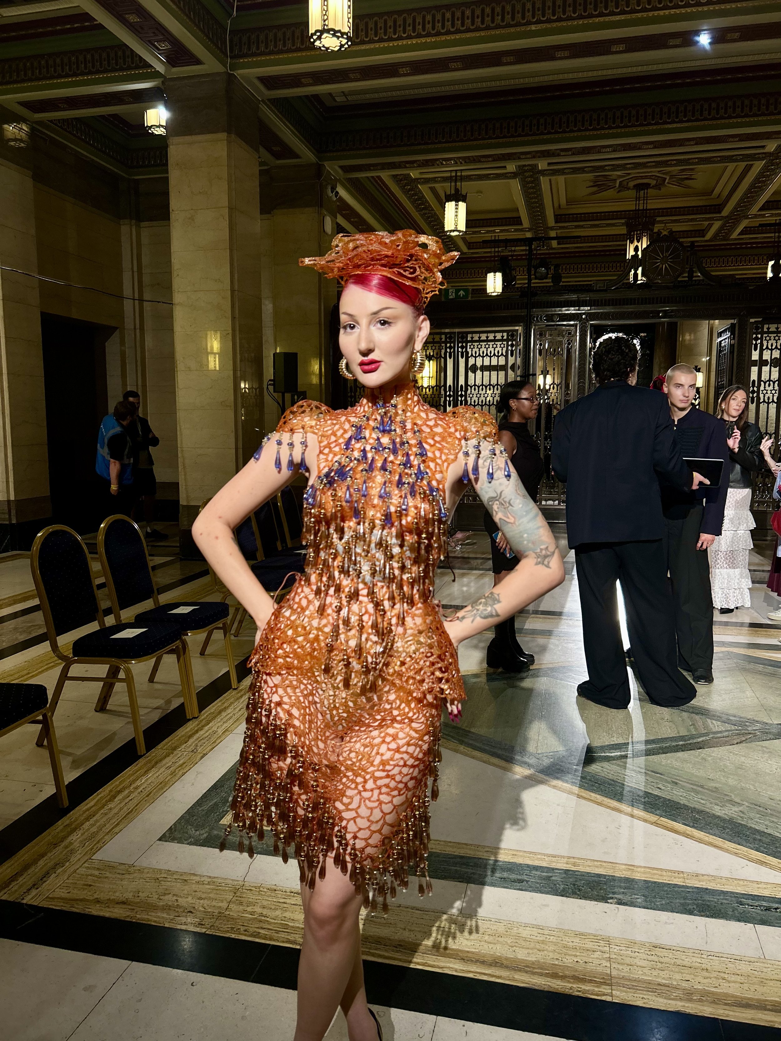 A woman with pink hair and tattoos wearing an orange and blue beaded dress with a matching headpiece standing in a decorated hall.