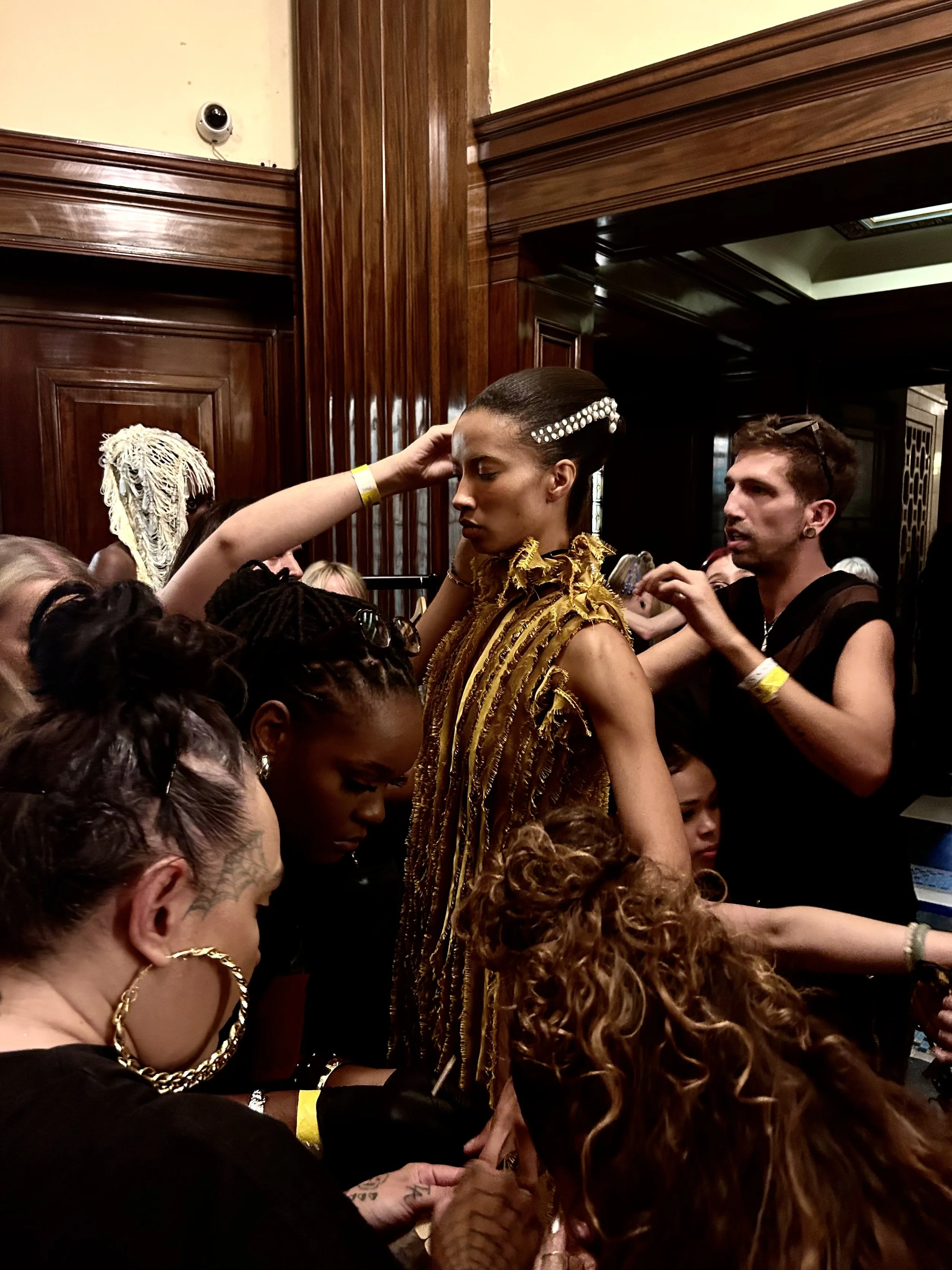 Models and makeup artists backstage at a fashion show, with one model in a gold, textured outfit and hair styled with pearls, surrounded by team preparing her.