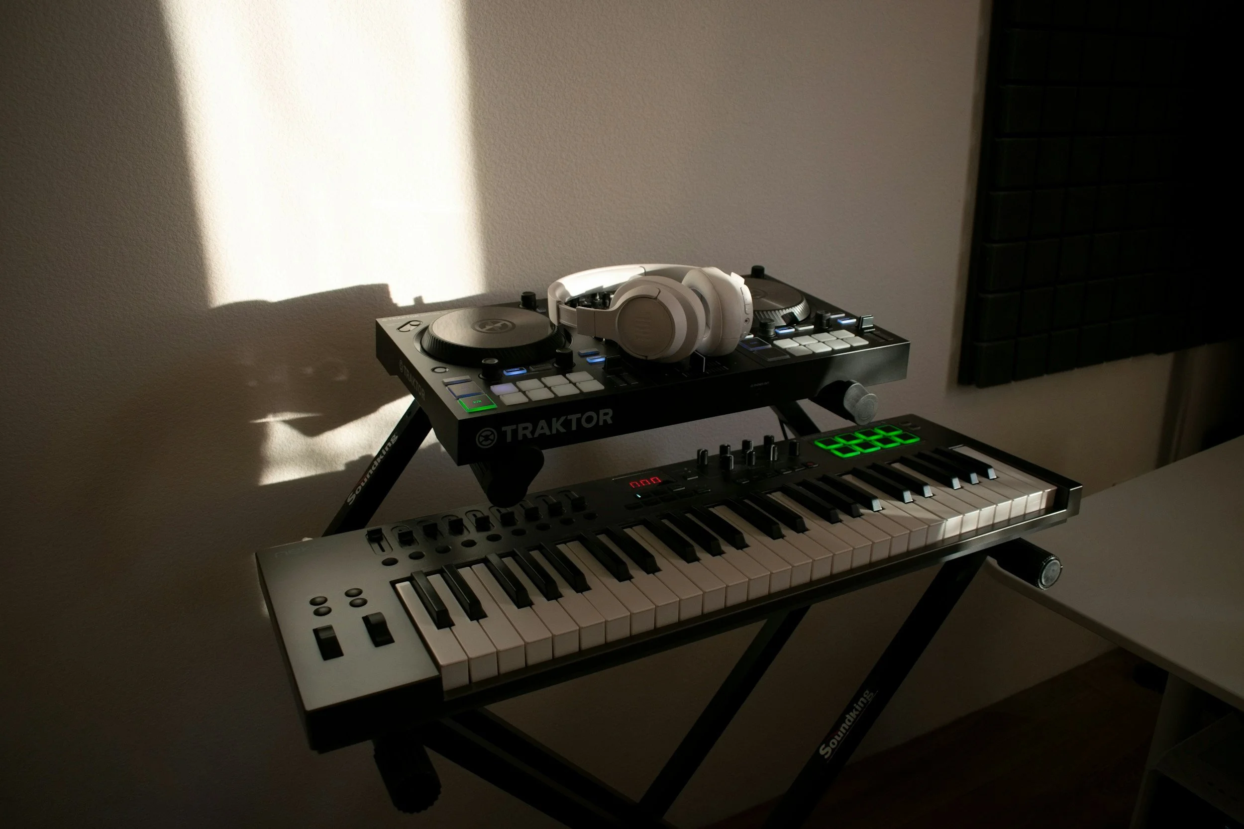 A music setup with a keyboard, a DJ controller, and white headphones on top of the DJ controller, all on a stand in front of a plain wall.