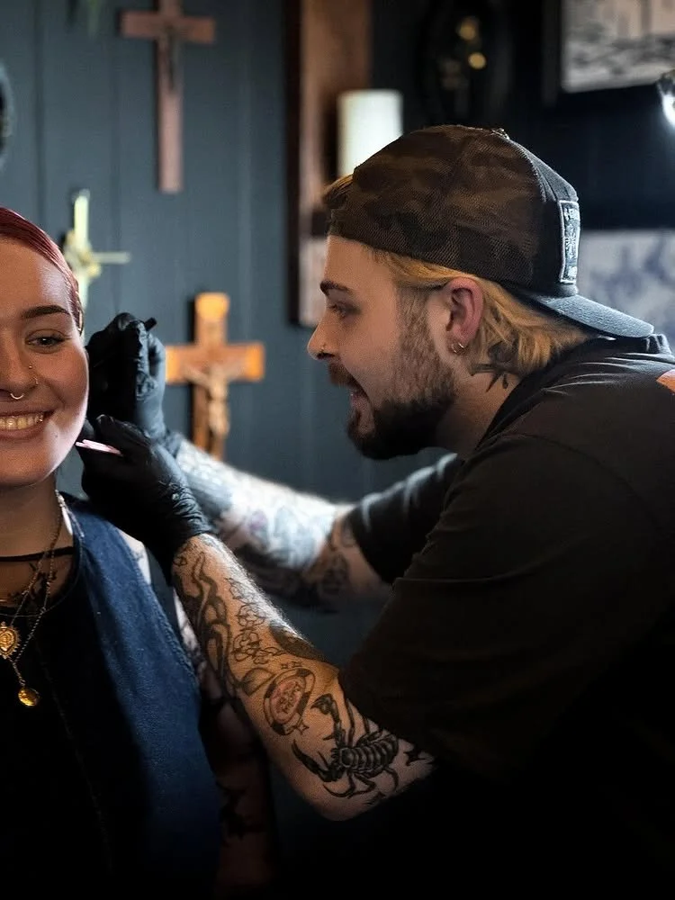 A person with tattoos on their arm and piercings getting a tattoo on their shoulder by a tattoo artist in a tattoo studio with Christian crosses in the background.