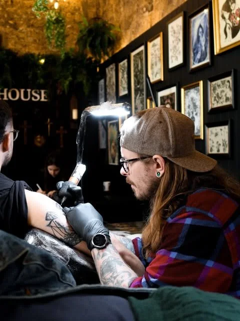 A tattoo artist in a plaid shirt and baseball cap tattooing a person's arm in an art gallery with framed pictures on the wall.