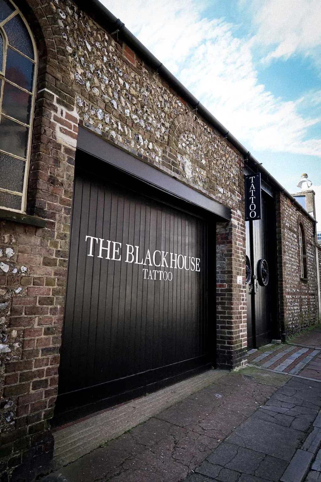 Exterior view of a tattoo studio named 'The Blackhouse'. The building has a brick wall facade with arched window and a black door with a sign for tattoo services.