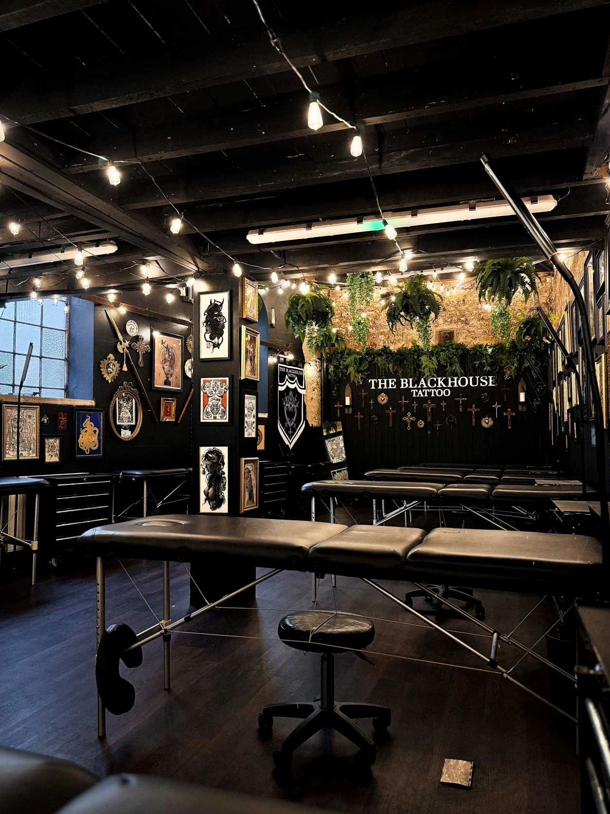 Tattoo studio with black walls decorated with artwork and framed prints, tattoo chairs, and hanging greenery with string lights overhead