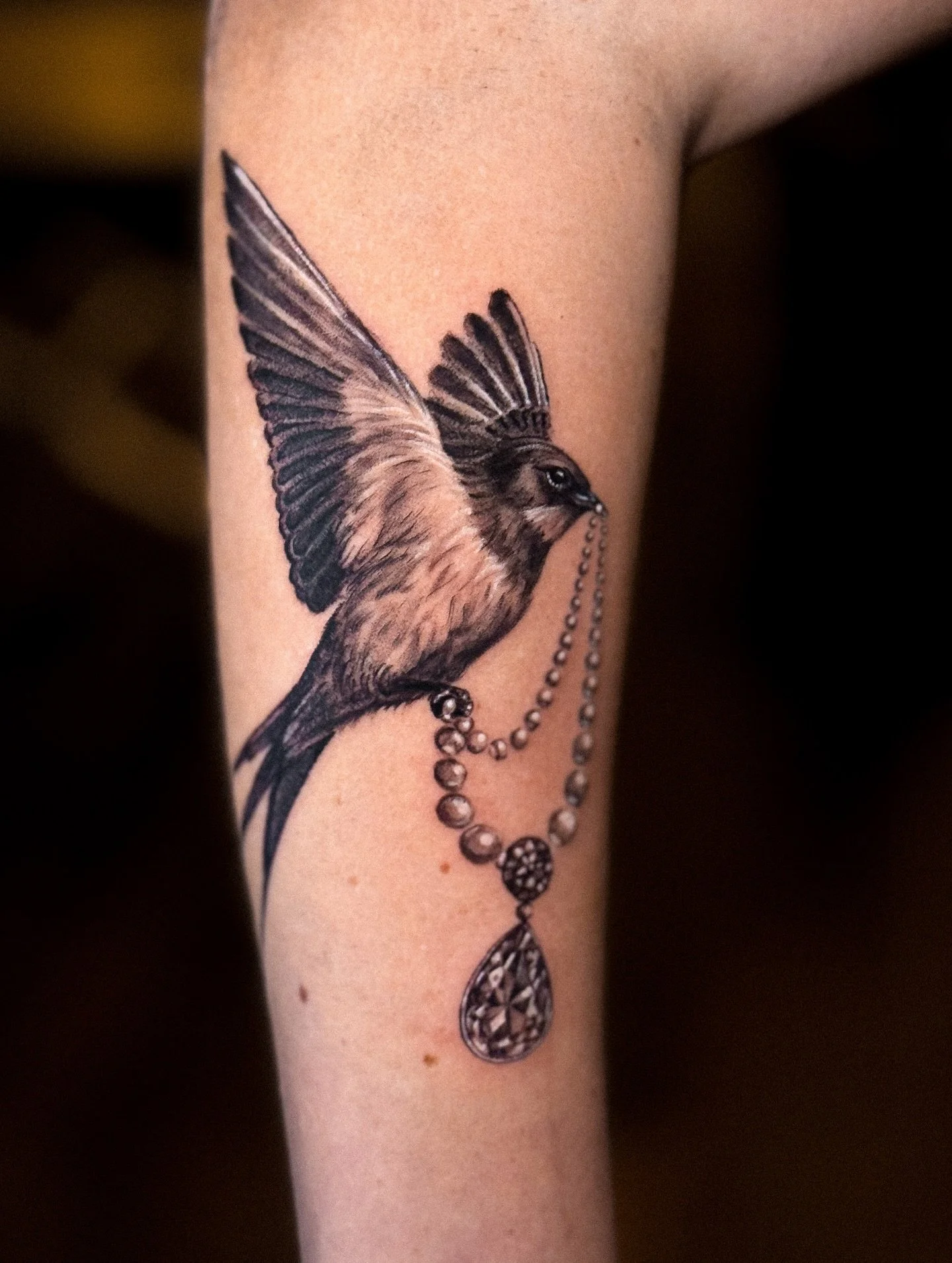 Tattoo of a bird with outstretched wings holding a bejeweled necklace with a large teardrop pendant on a person's arm.