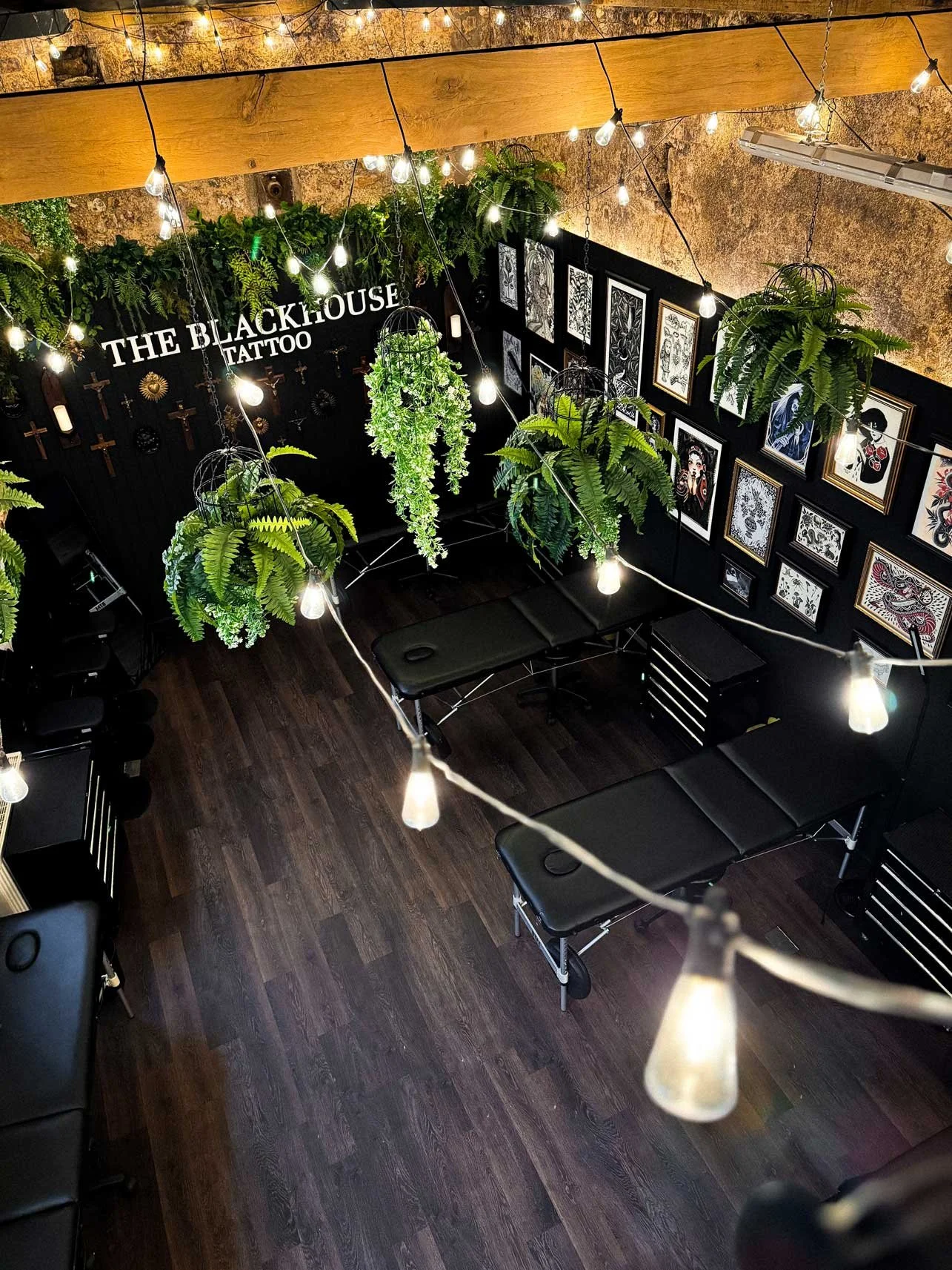 Interior of a tattoo studio named "The Blackhouse Tattoo" featuring black walls adorned with framed artwork, hanging green plants, and string lights overhead, with massage tables and various tattoo equipment.