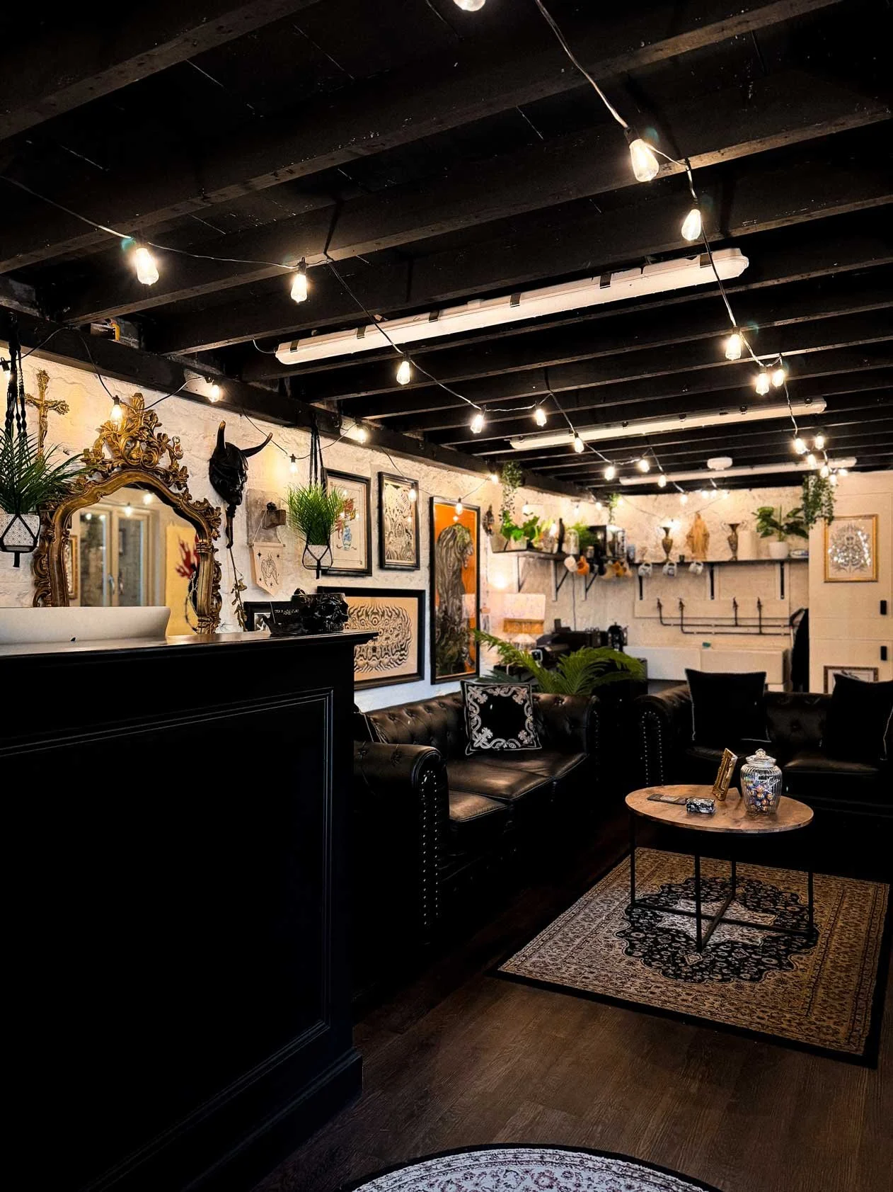 Cozy living room with string lights, black leather sofa, rug, wooden coffee table with decorative items, art and plants on the walls, and a shelf with vases and decor in the background.