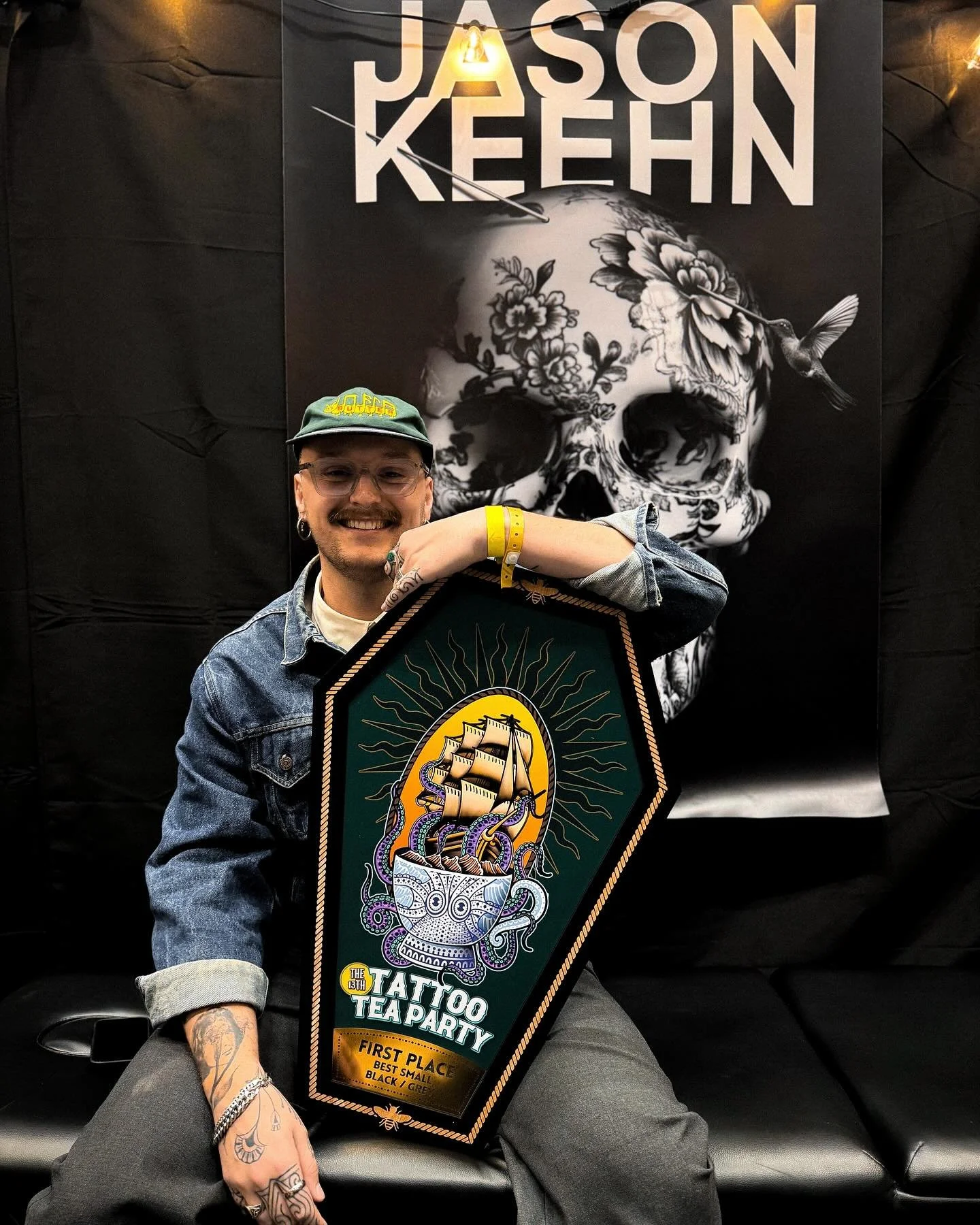 A man smiling and holding a large tattoo convention award plaque, sitting in front of a black background with a skull artwork and the name Jason Keehn.