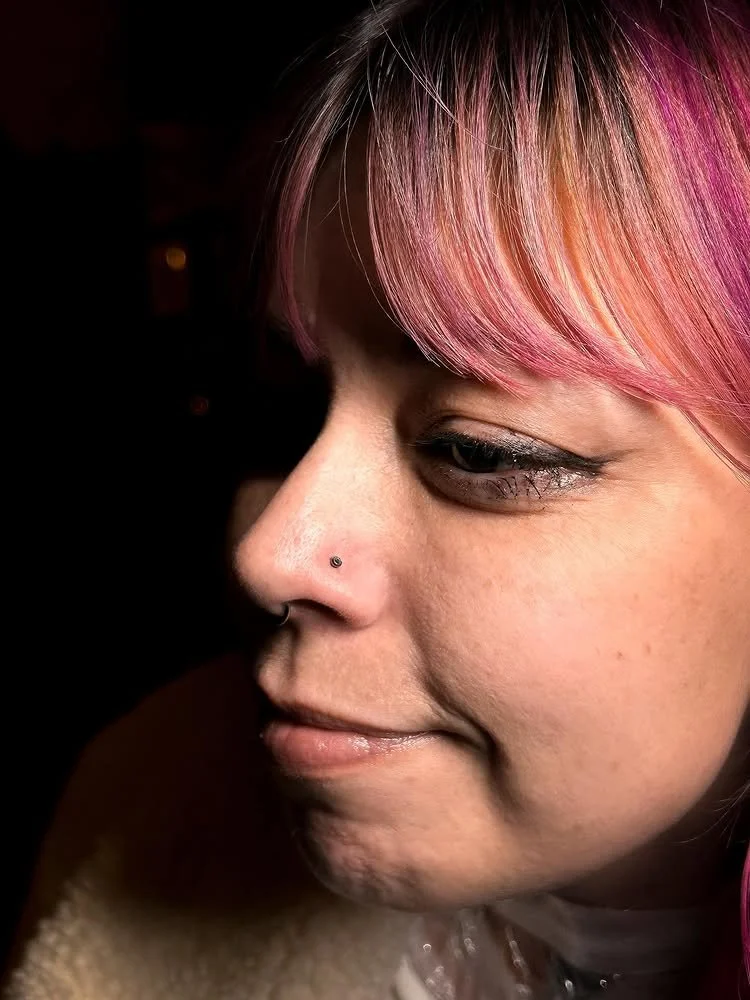 Close-up of a woman with pink hair, a nose piercing, and wearing black makeup around her eyes, looking downward with a slight smile.
