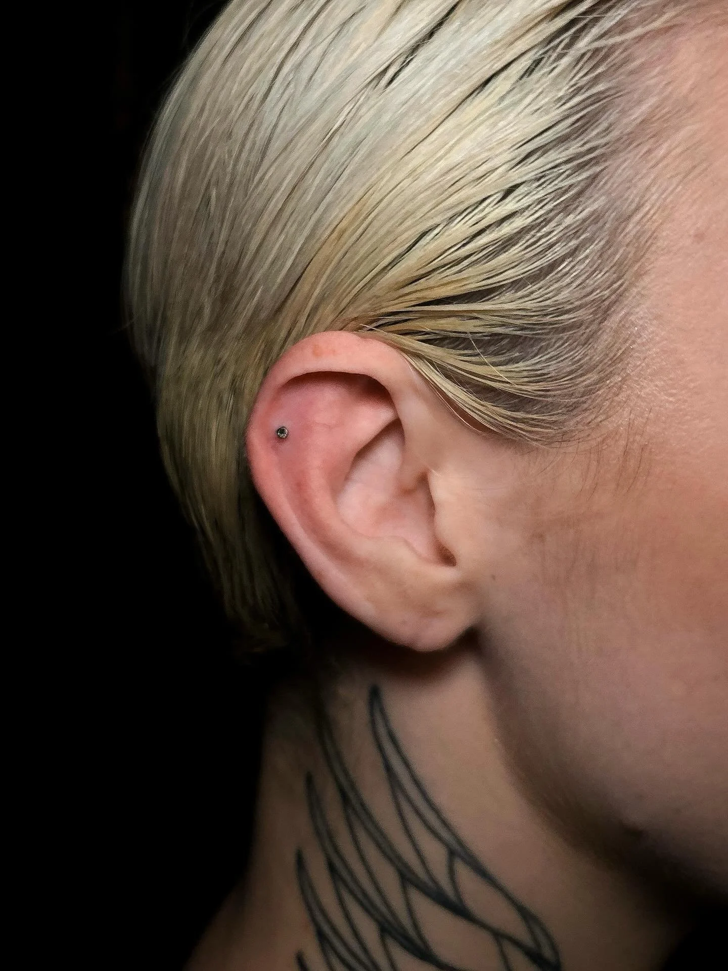 Close-up of a person's ear with a small piercing, part of their blond hair, and a tattoo on their neck.