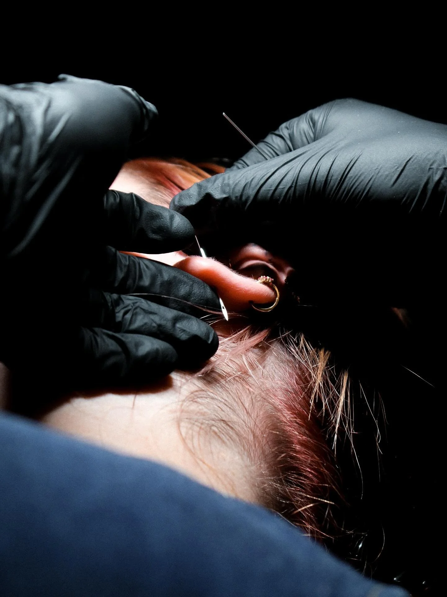A person receiving a piercing on their ear, with black-gloved hands holding the ear and inserting a piercing needle.