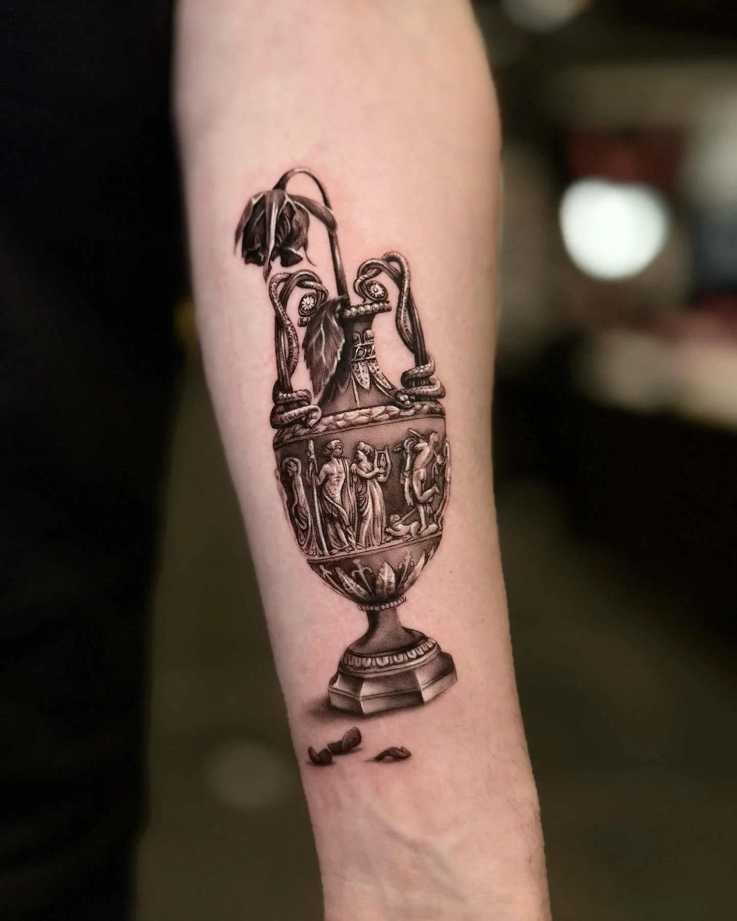 Tattoo of an ornate ancient Greek or Roman-style urn with decorative reliefs and figures, with coffee beans below it.