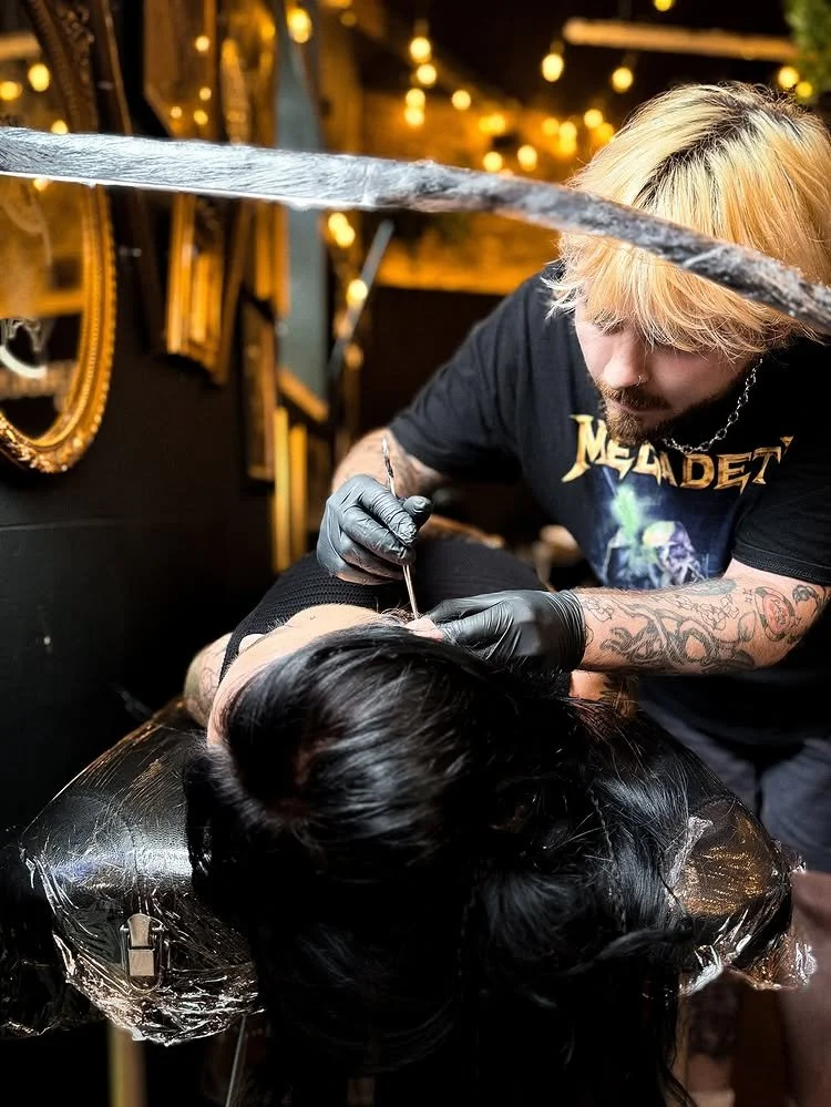 A tattoo artist with blond hair and tattoos on his arms tattooing a woman with long dark hair lying on a black chair.