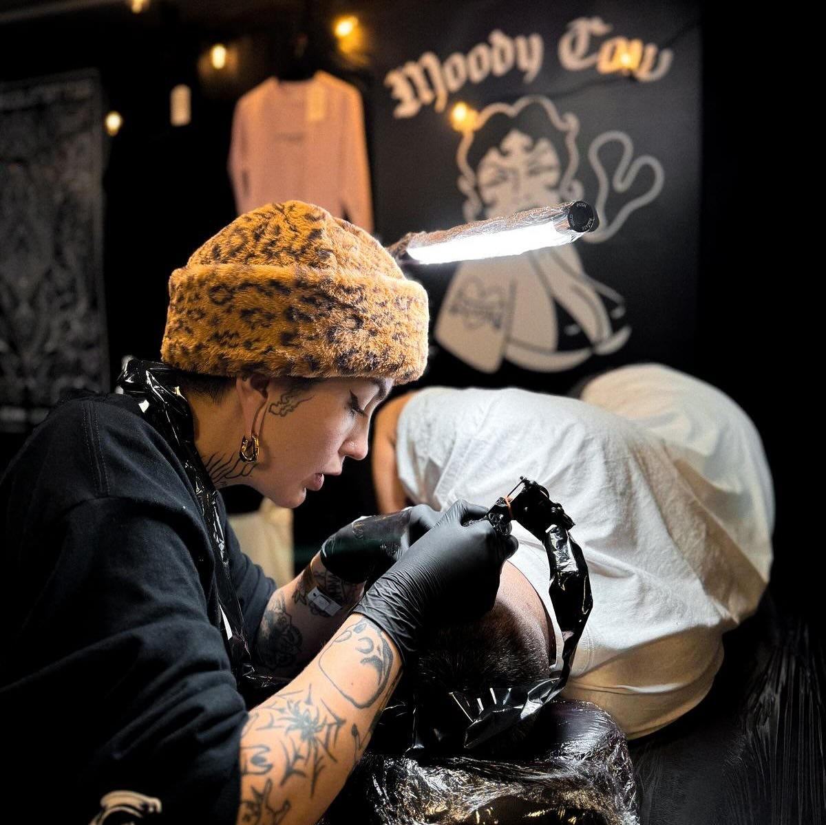 Tattoo artist with leopard print hat working on a client's head inside a tattoo parlor, with Mooby Cat logo on the wall behind.