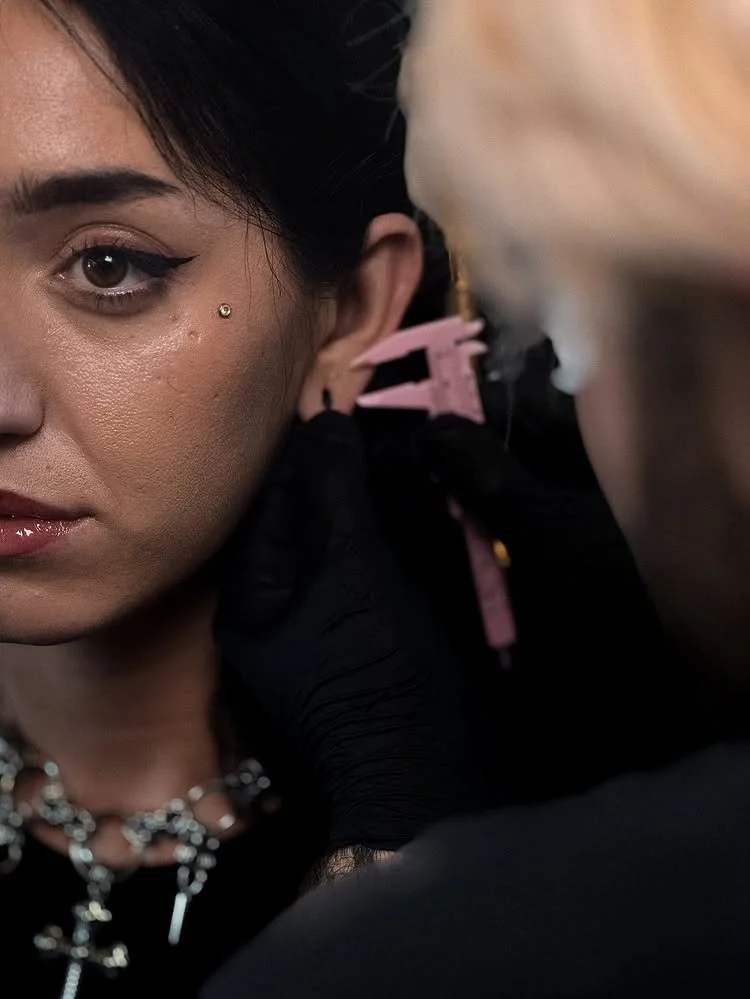 A woman getting a piercing on her ear from a person wearing black gloves. The woman has makeup and jewelry, including a necklace and a nose piercing.