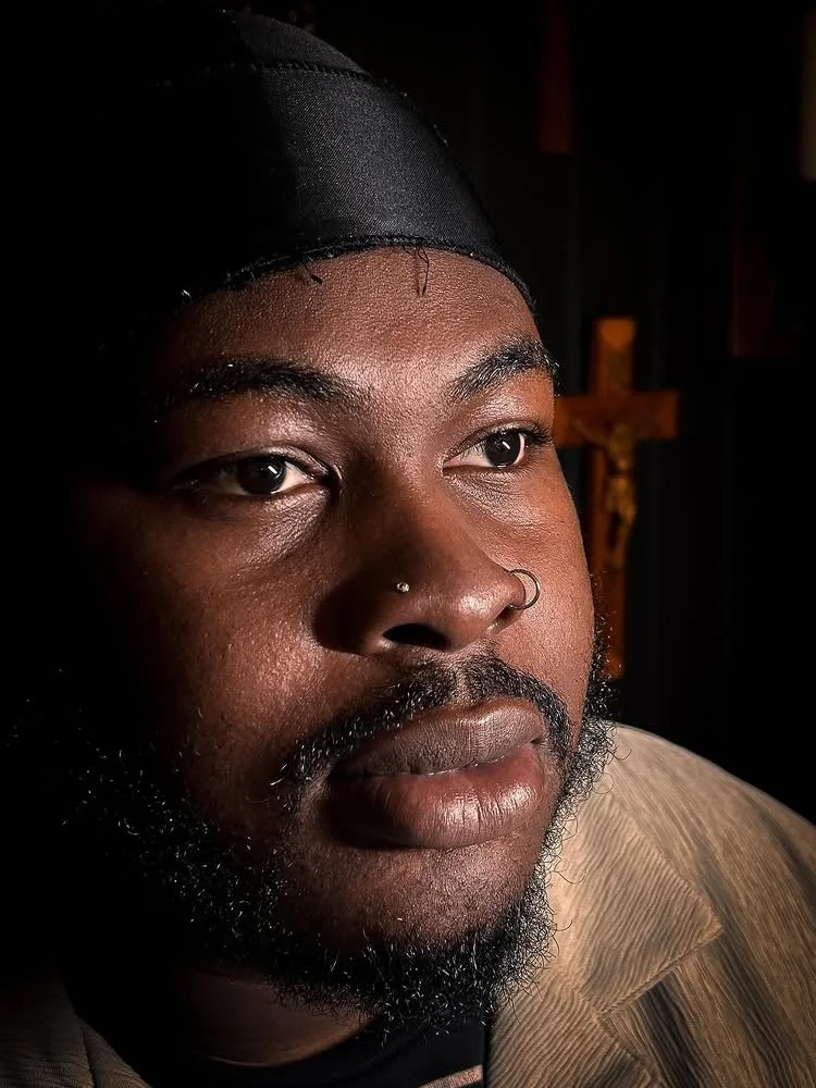 Close-up portrait of a Black man wearing a black head covering, with a nose and septum piercing, and a beard, in front of a wooden cross with a figure of Jesus in the background.
