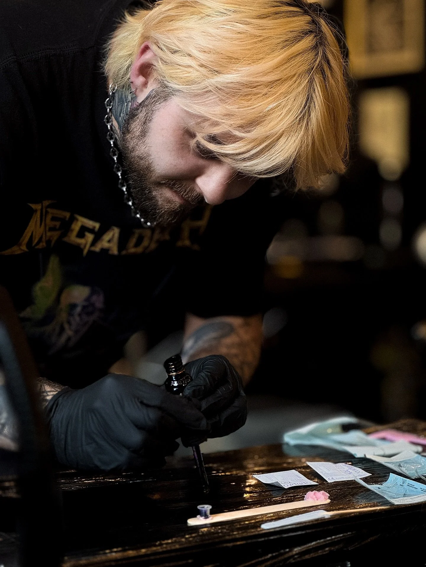 A person with blond hair, beard, and tattoos, wearing black gloves, is carefully applying ink to a tattoo on a person's skin, with tattoo-related supplies nearby.