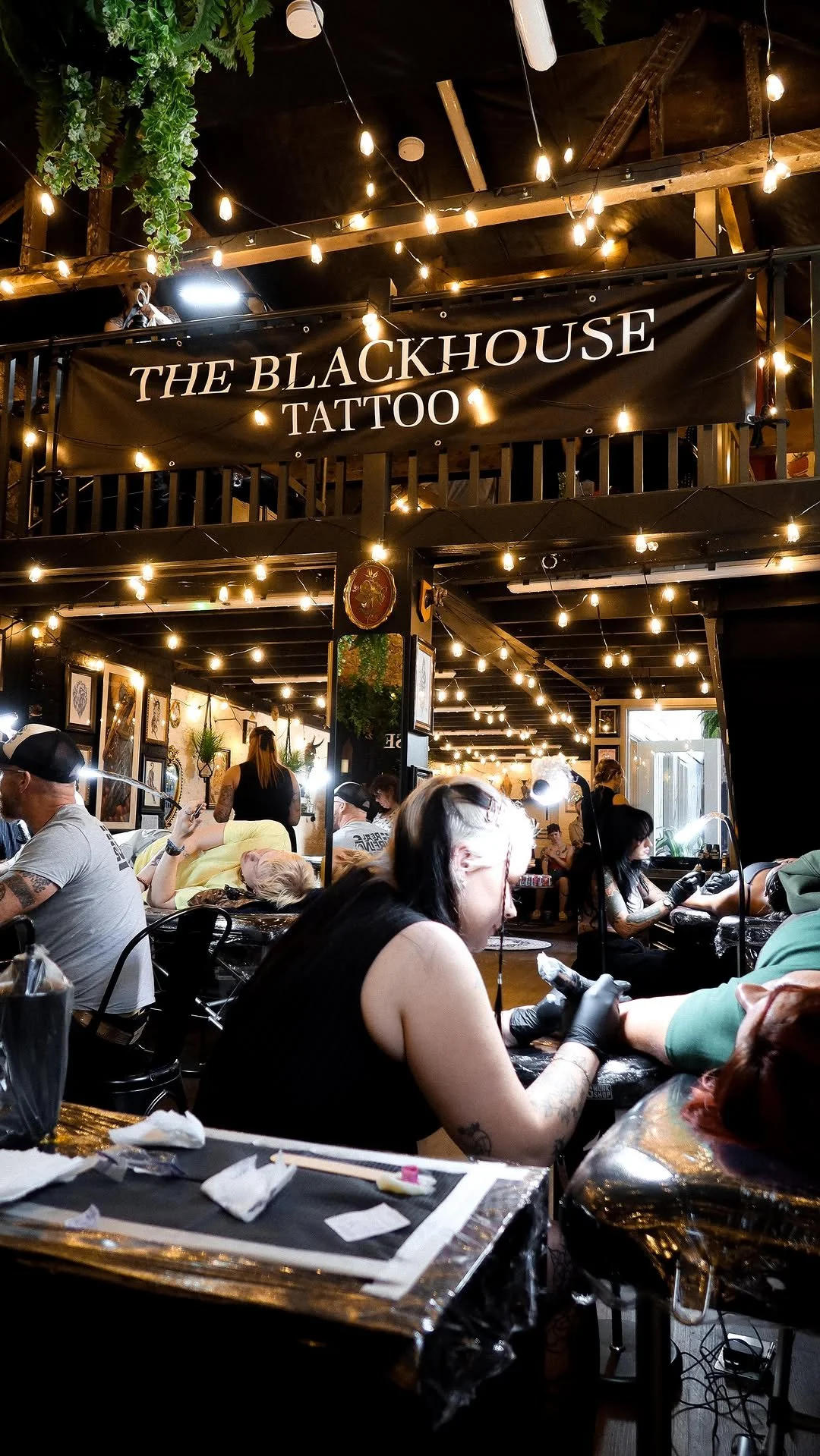 Tattoo artists tattooing clients at The Blackhouse Tattoo studio, illuminated by string lights inside a bustling, artistic tattoo shop.