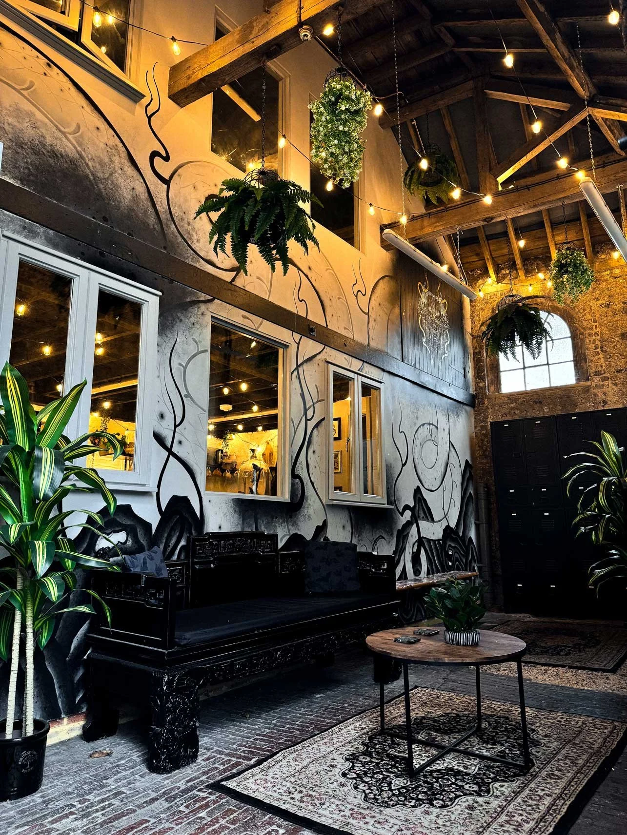 Indoor loft space with industrial design, string lights, hanging plants, black wall mural, large windows, black wooden bench, and round wooden table with potted plants.