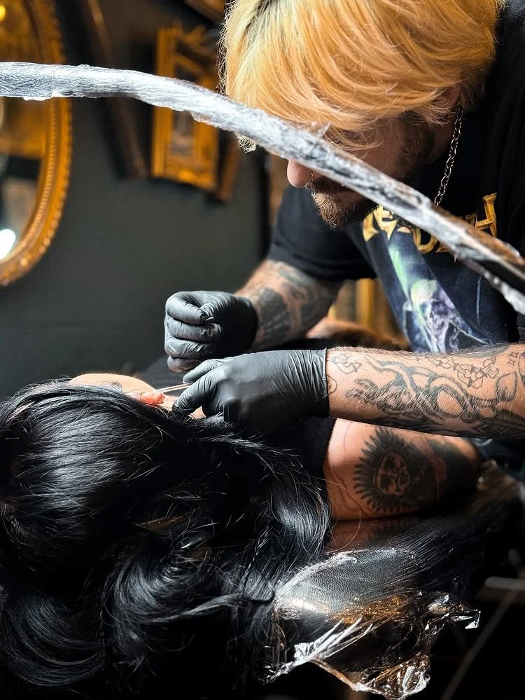 Tattoo artist with blonde hair and tattoos, wearing black gloves, working on a woman's hair in a salon with dark walls and gold-framed mirrors.