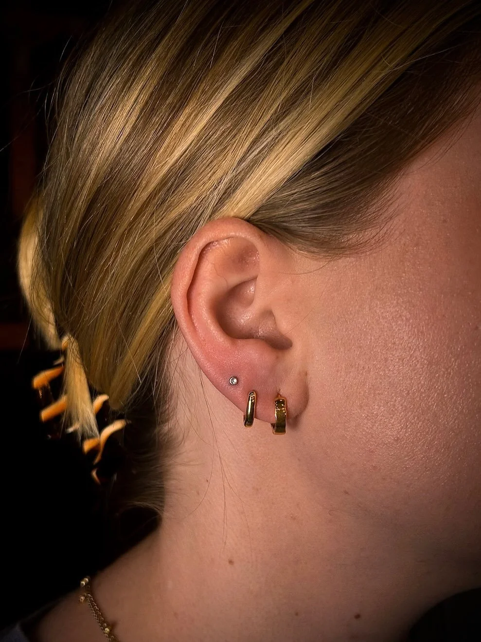 Close-up of a person's ear with three earrings: a small stud, a thin hoop, and a thicker hoop, with blonde hair tucked behind the ear and a portion of the face visible, showing skin and a necklace.