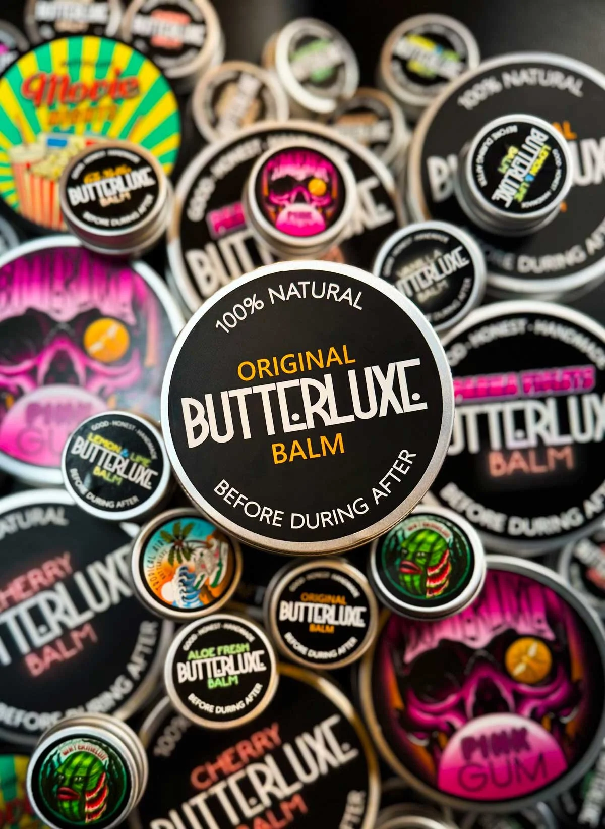 A collection of Butter Luxe balm containers with colorful, tropical-themed and skull-themed labels, prominently featuring a black lid with white and orange text that says '100% Natural Original Butter Luxe Balm Before During After'.