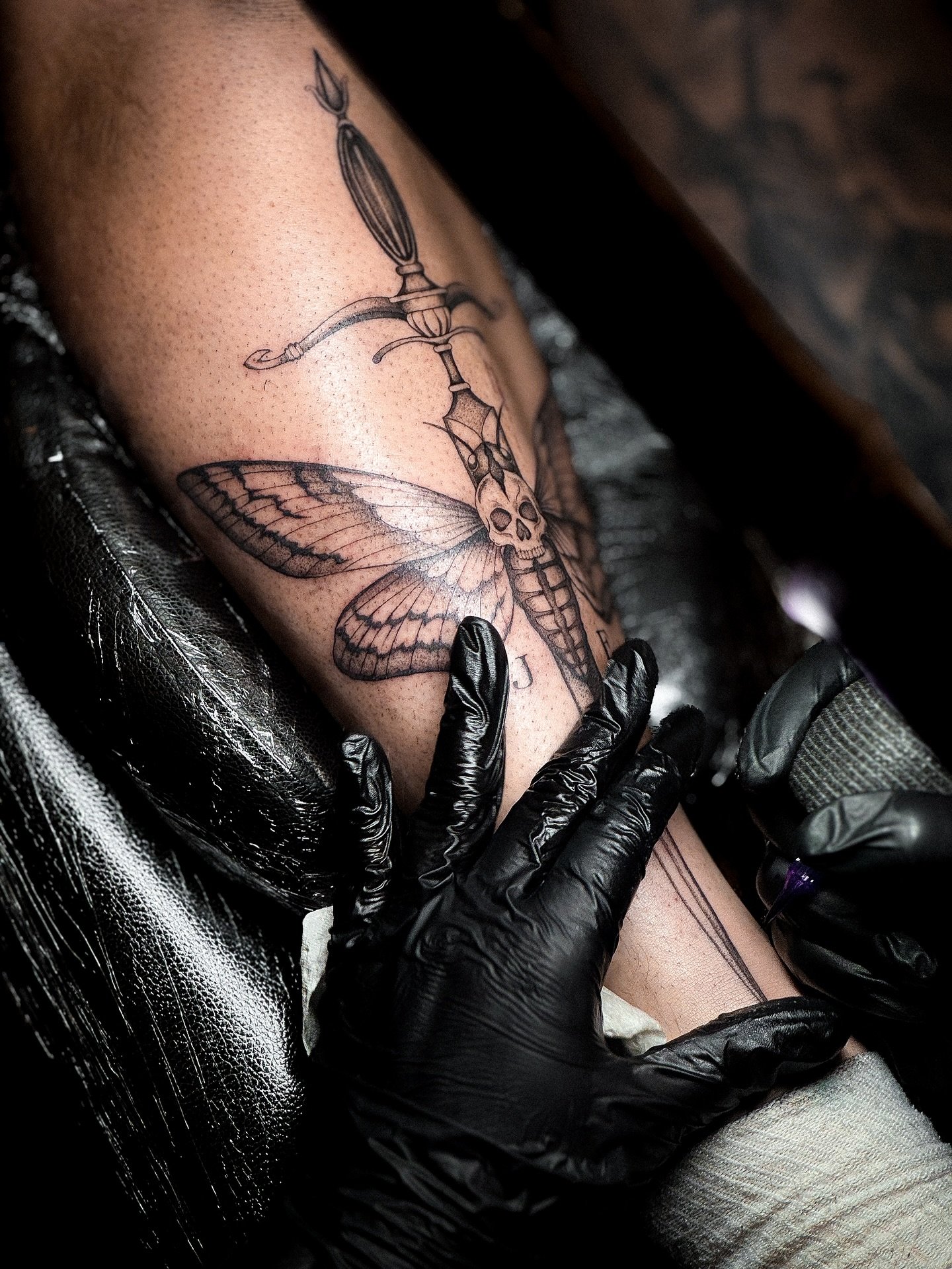 A tattoo artist in black gloves is creating a tattoo of a moth with a skull on its thorax, along with other detailed designs, on a person's forearm.