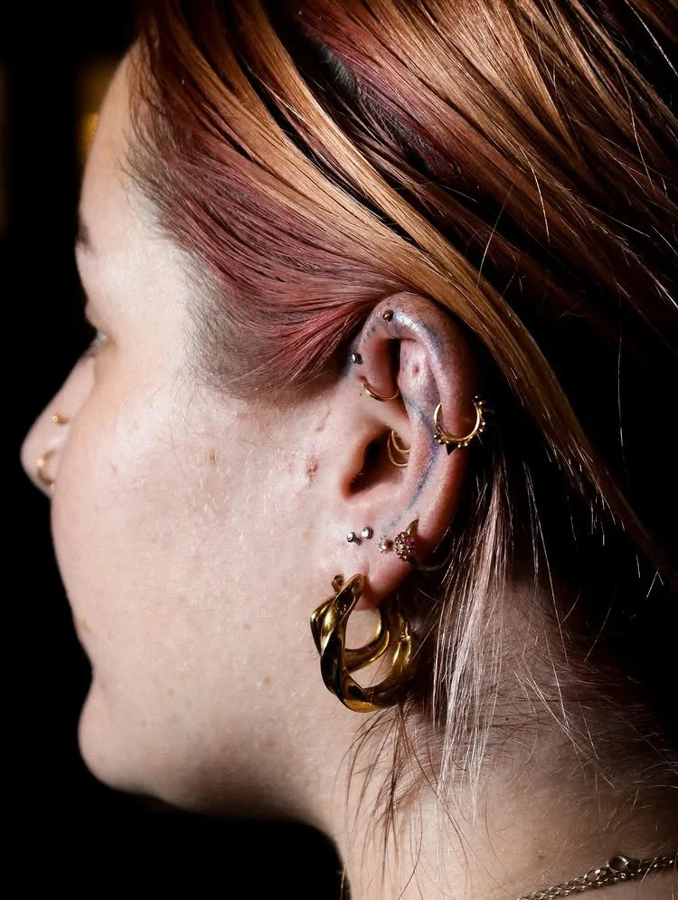 Close-up of a woman's ear with multiple earrings, including gold hoops, studs, and a budding cartilage piercing, with reddish-brown hair partially covering her face.