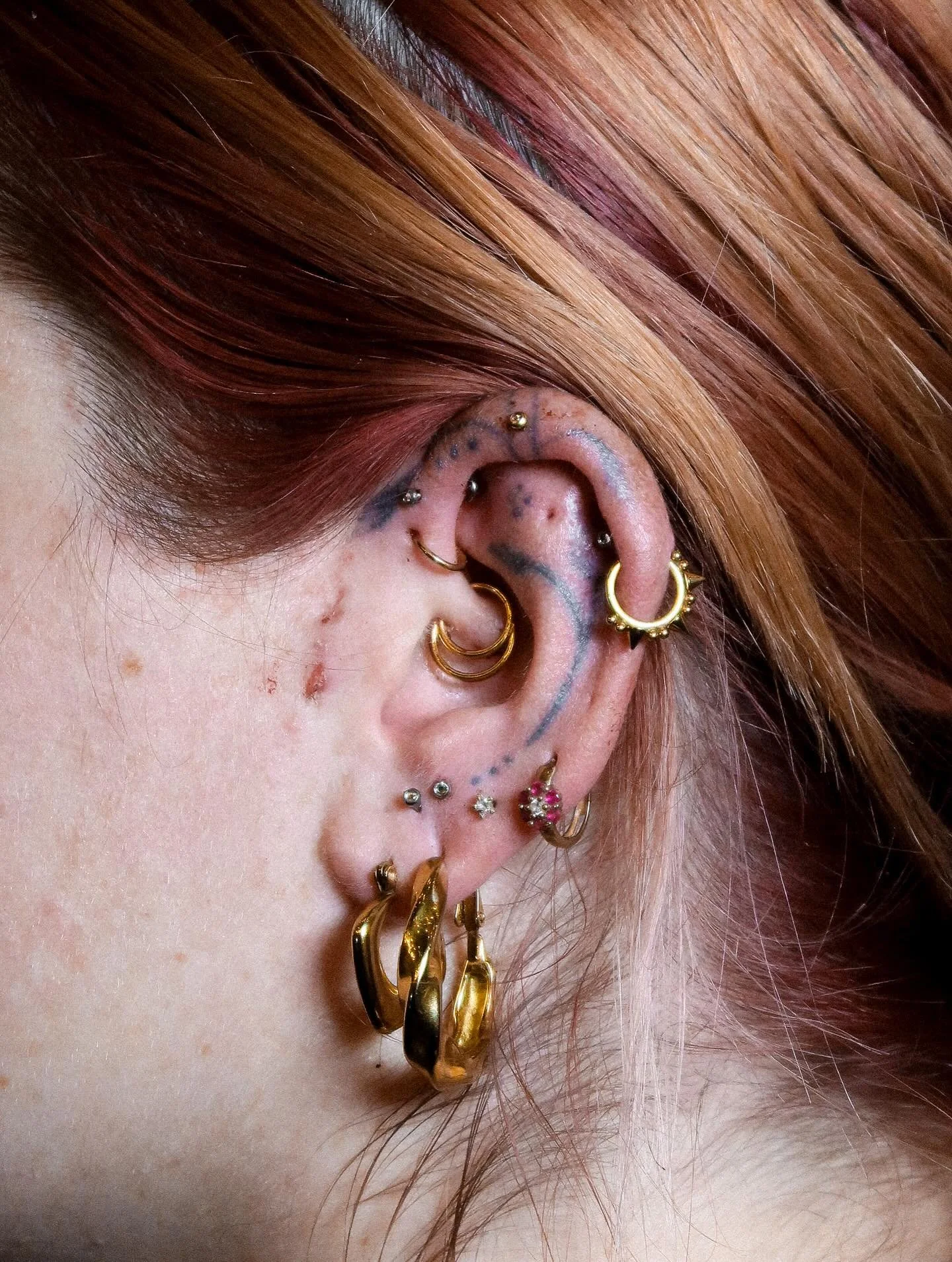 Close-up of a person's ear with various gold earrings, tattoos, and colored hair.
