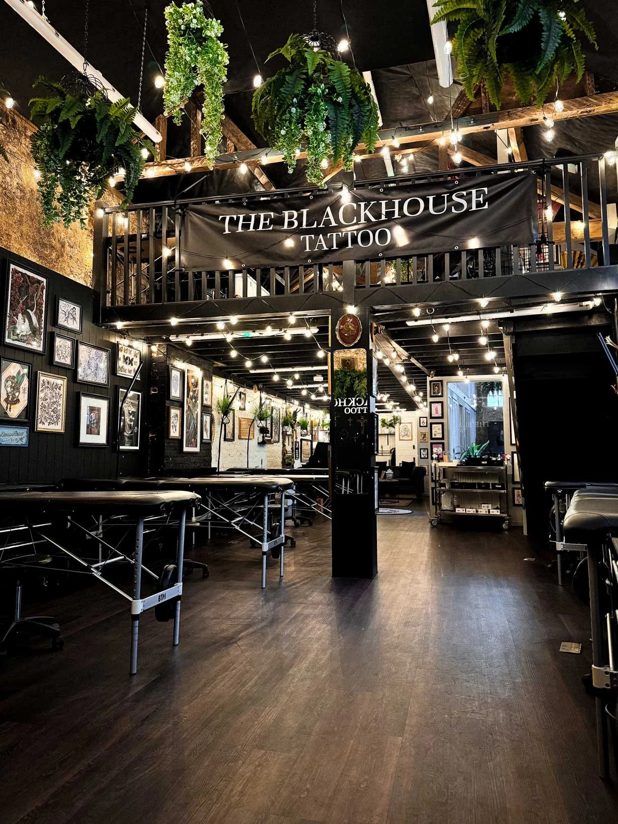 Interior of a tattoo studio called 'The Blackhouse Tattoo' with framed artwork on the walls, hanging plants, and tattoo beds for clients.