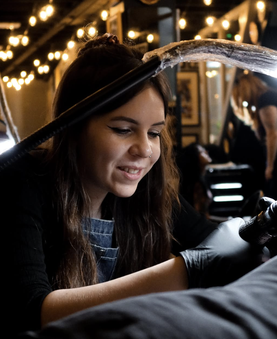 A young woman with long brown hair, wearing a black shirt and denim apron, tattooed arm, is smiling and getting a tattoo on her arm from a tattoo artist in a dimly lit room decorated with warm string lights.