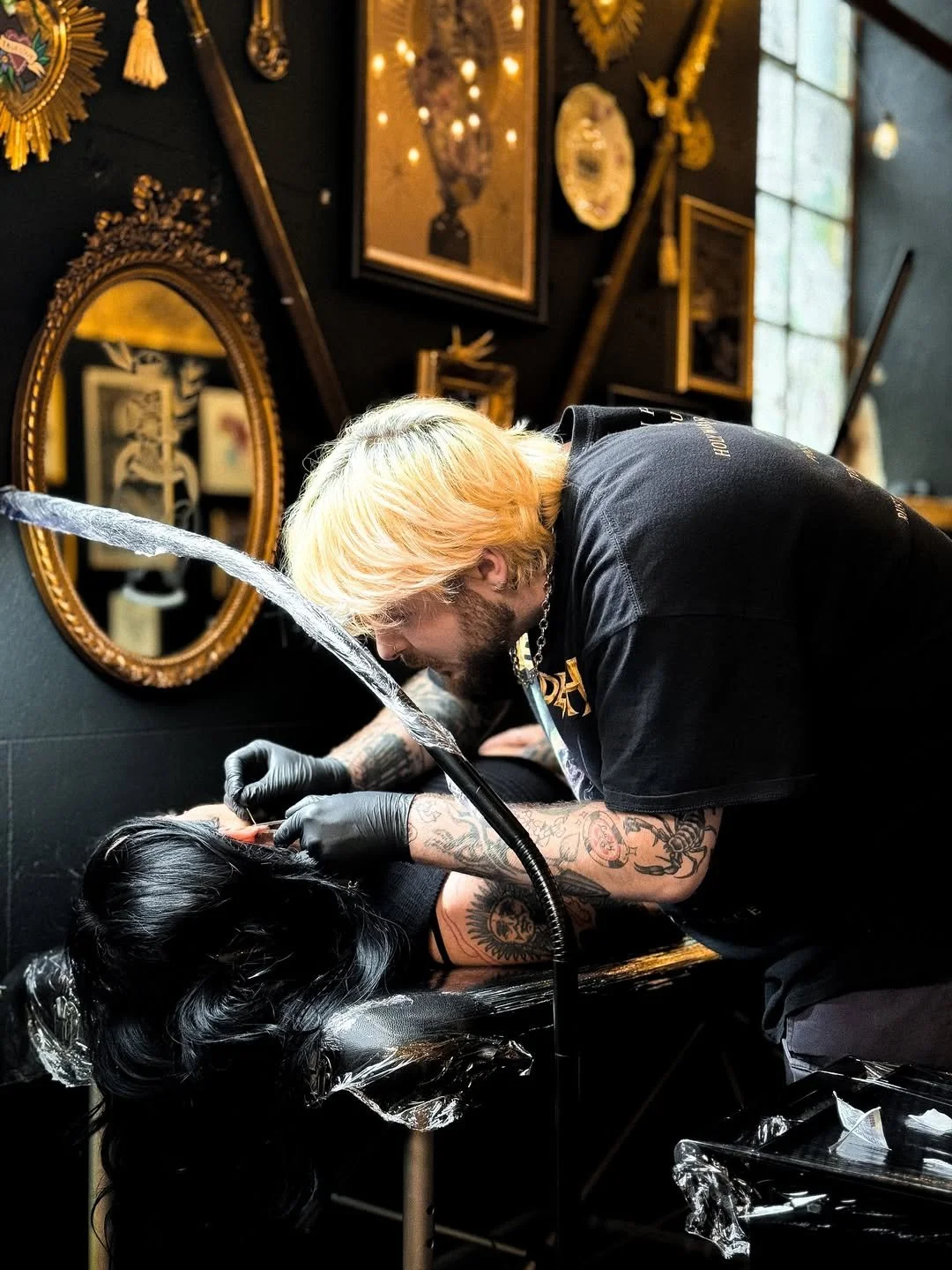 A tattoo artist with blonde hair and tattoos on his arms is tattooing a person's arm in a tattoo studio. The studio has dark walls, framed artwork, and gold decorative mirrors.