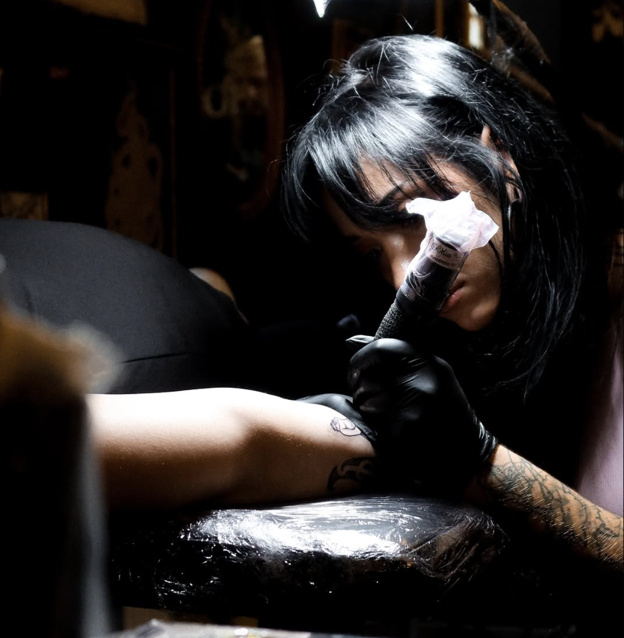 A tattoo artist with black hair and tattoos on her left arm is tattooing a person's arm in a dimly lit tattoo studio.