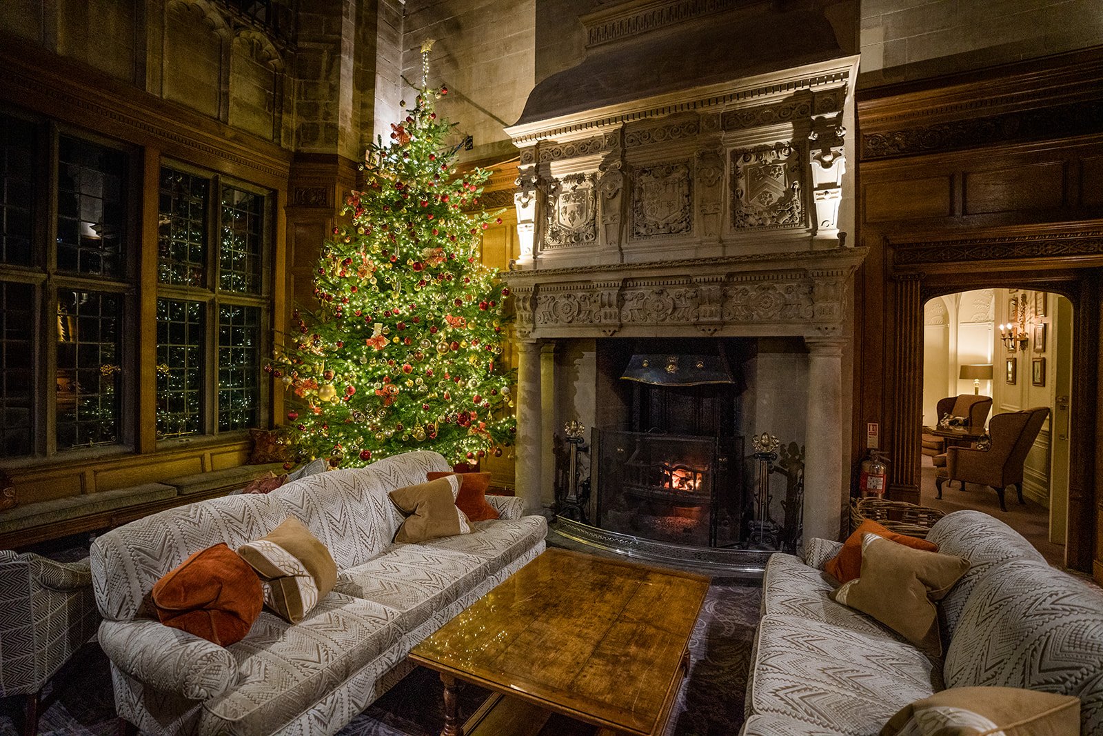 A picture of the sitting room with a grand fireplace, a gigantic Christmas tree with red baubles and lights.