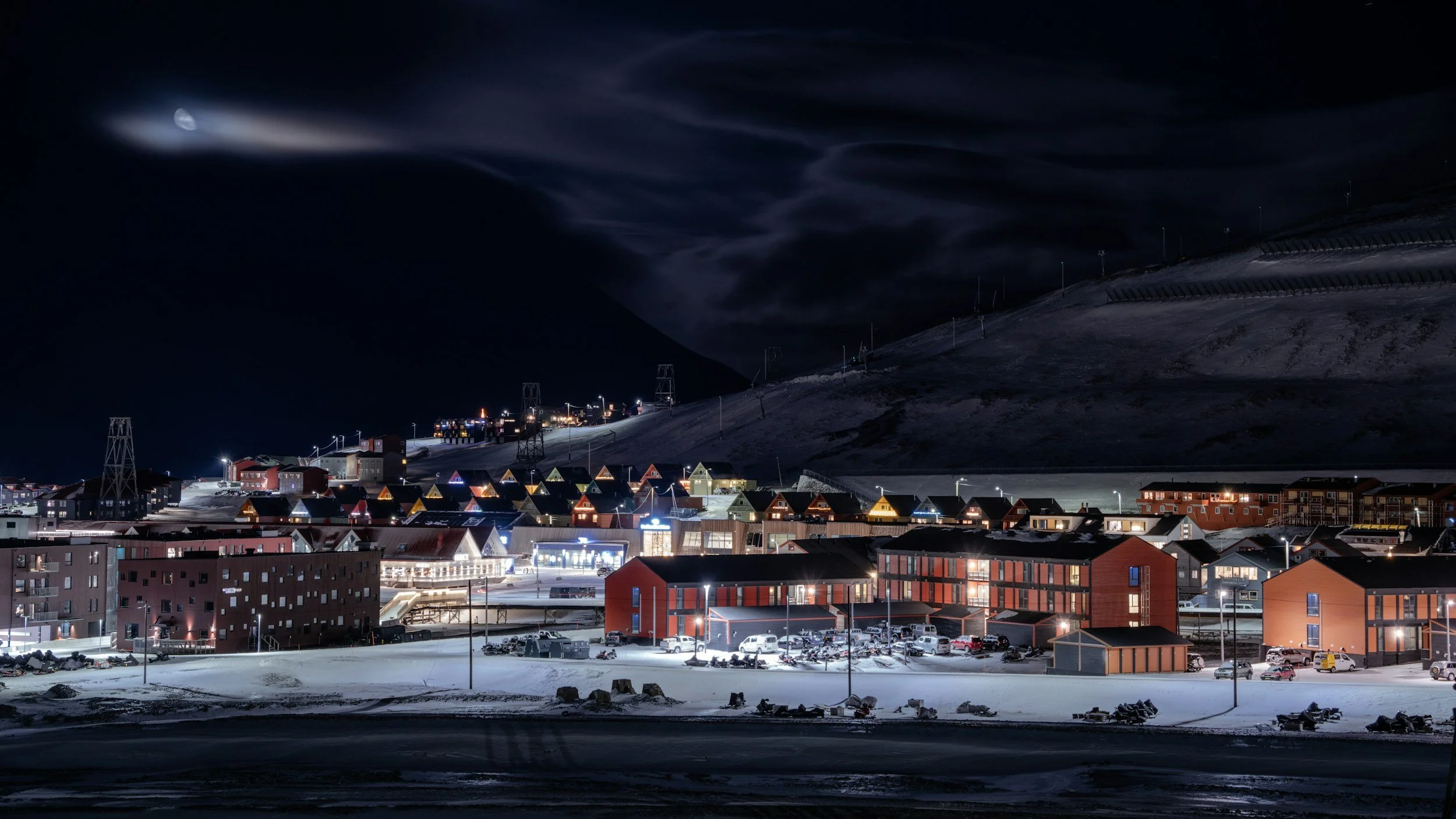 Night view of Longyearbyen in the polar night, with glowing houses, snow-covered ground and a dramatic Arctic sky.