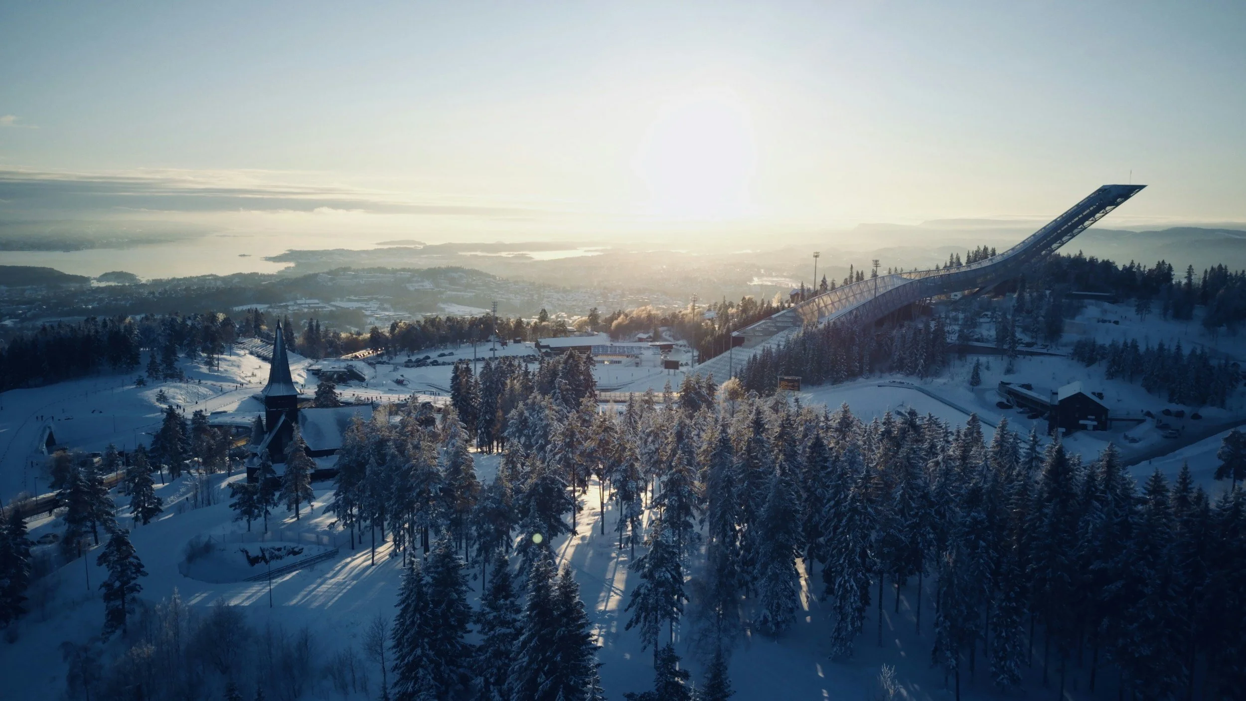 Winter view from Holmenkollen overlooking Oslo, showing snow-covered forest, ski facilities and the city under low winter light.
