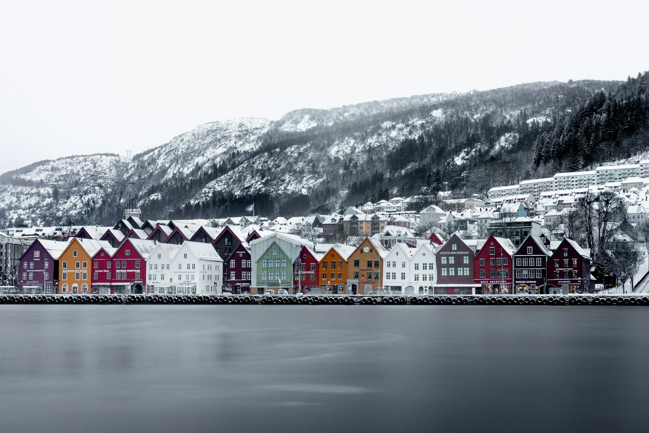 Winter view of Bryggen showing colourful wooden houses along the harbour with snow-covered hills in the background.