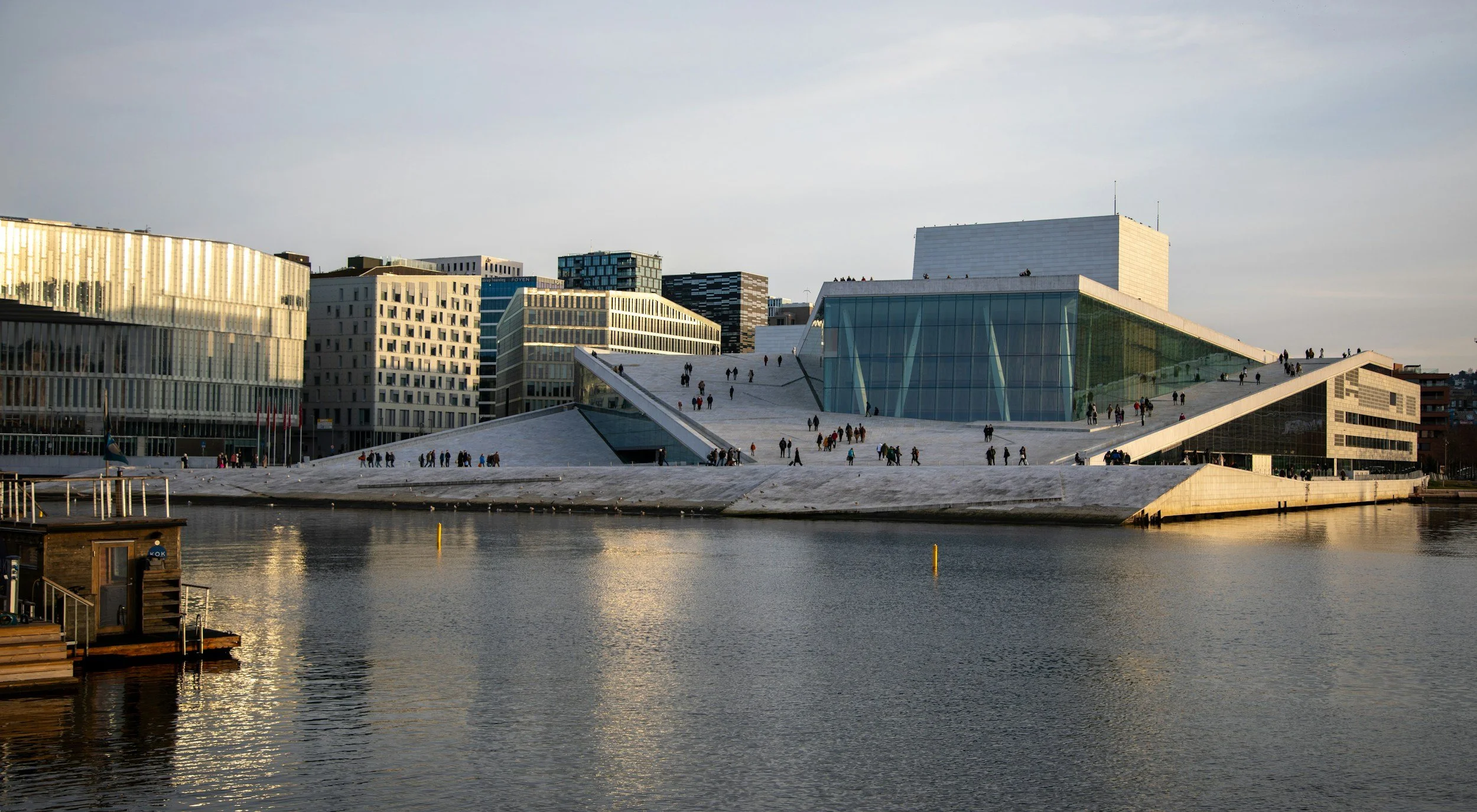 A picture of the Opera House in Oslo at sunset in winter. People are walking at the top of the Opera house, and the Opera house itself has a thin layer of snow on it. The buildings in the background is lit up by the disappearing sun.