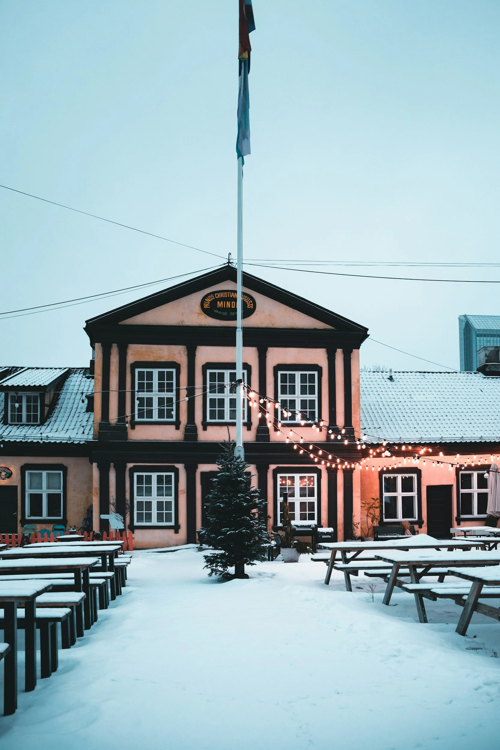 A picture outside a restaurant in Oslo during winter, the benches outside are covered in snow, the christmas tree is lit up.