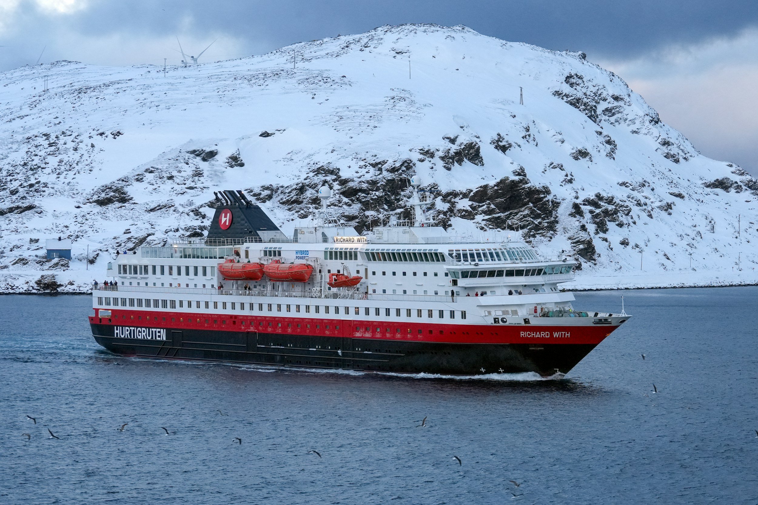 The Hurtigruten ship sailing in the fjord in front of snow covered mountains.