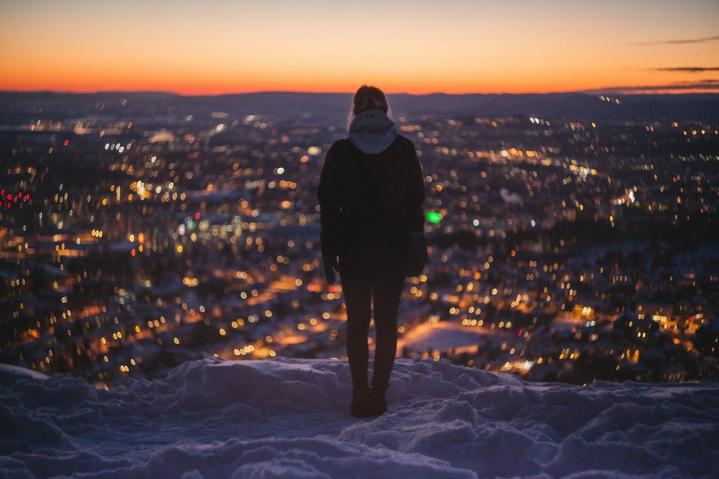 A woman standing in snow at sunset looking over the lights from the city at the viewpoint in Grefsenkollen, Oslo.