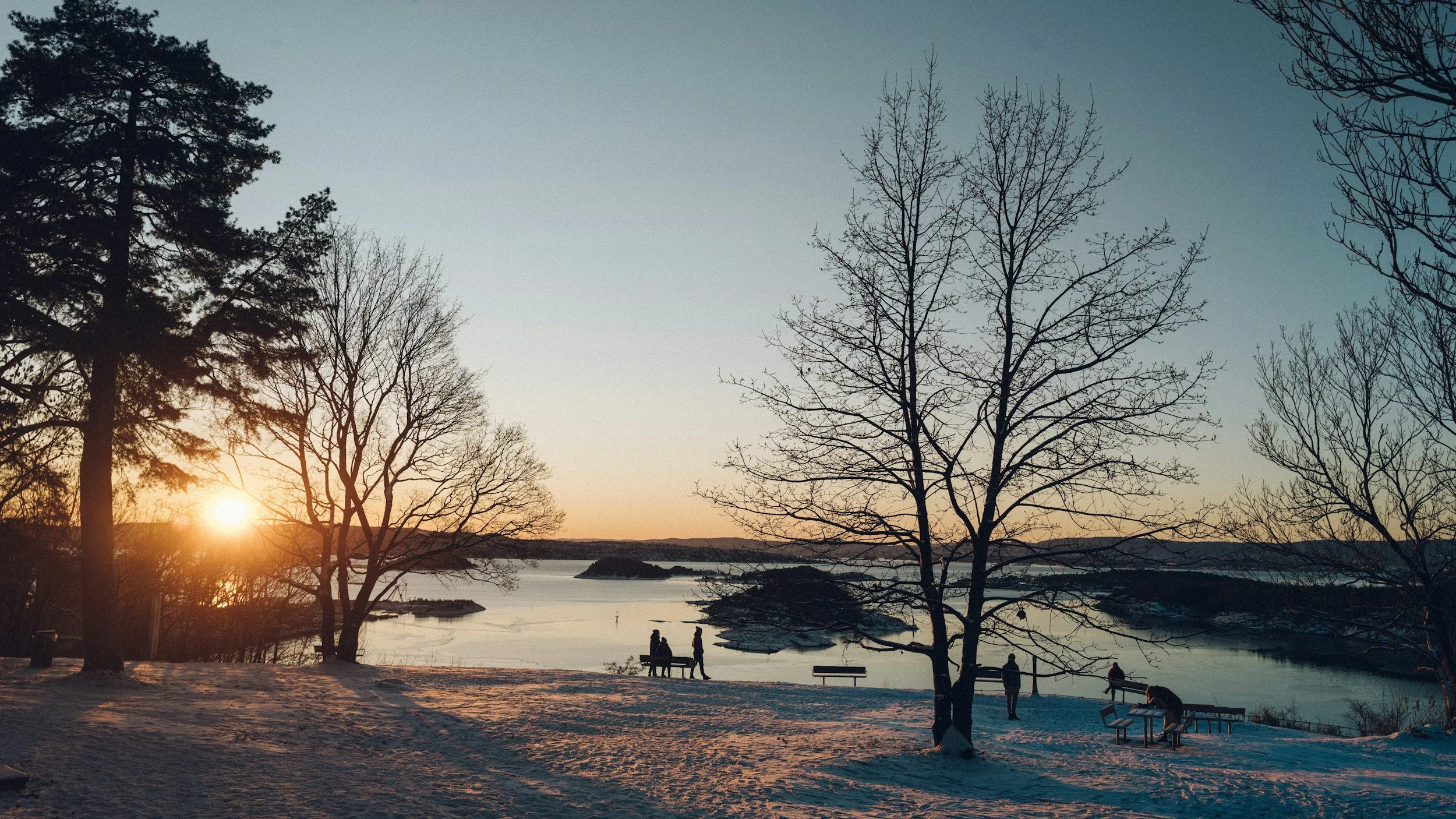 A picture from the viewpoint at Ekebergparken in Oslo, people are sitting at benches and walking around, enjoying the view of the sunset over the Oslo fjord. The ground is covered in snow.