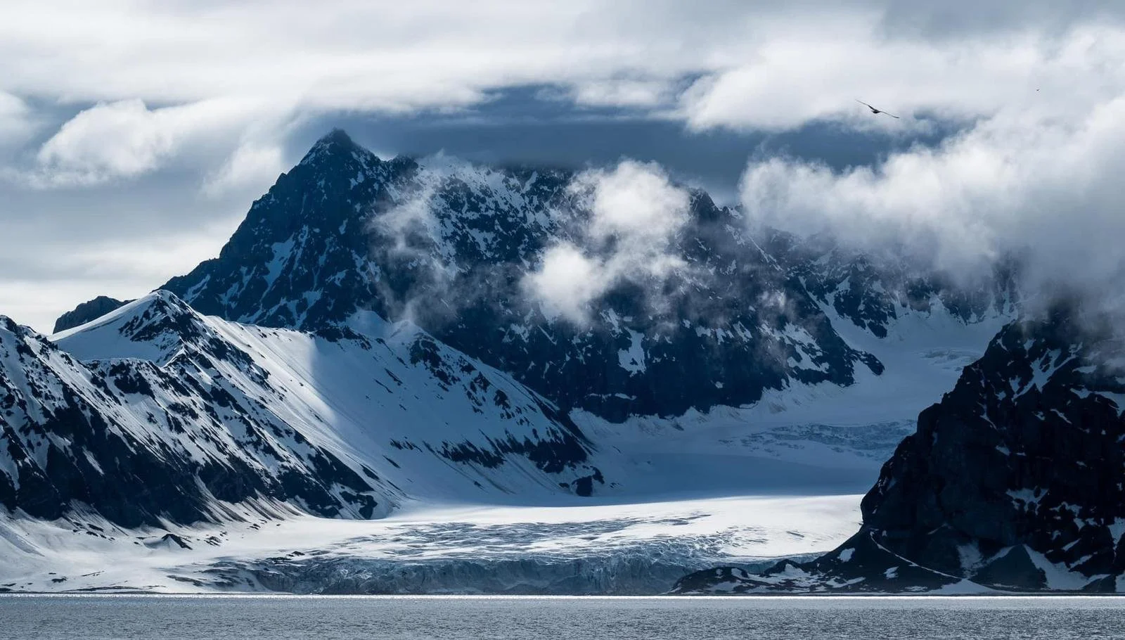 Dramatic Arctic landscape in Svalbard with towering snow-covered peaks, drifting clouds and a glacier meeting the fjord.