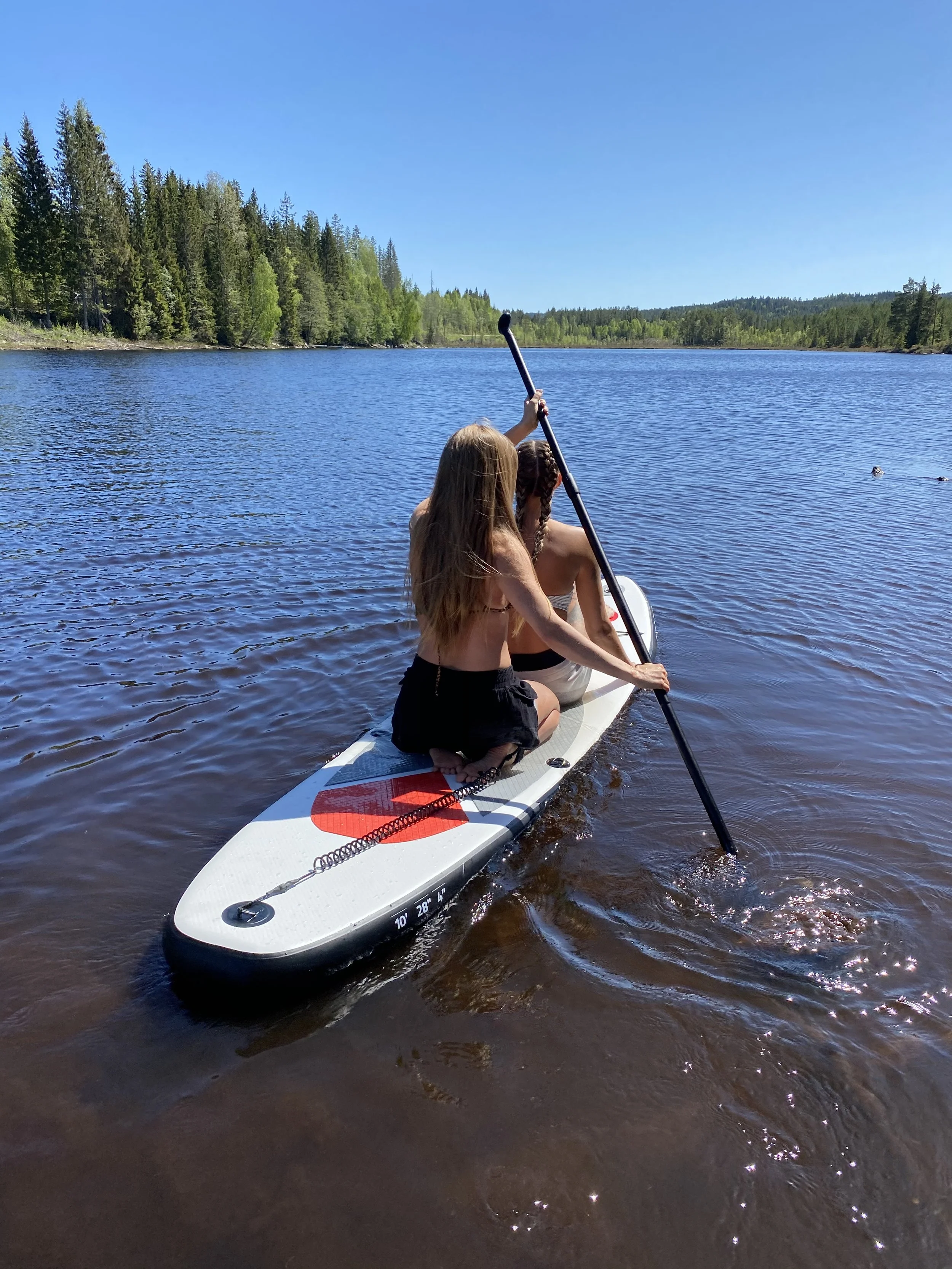 Paddleboarding on a calm lake in Norway during a warm summer day, wearing light summer clothing suitable for swimming and water activities.