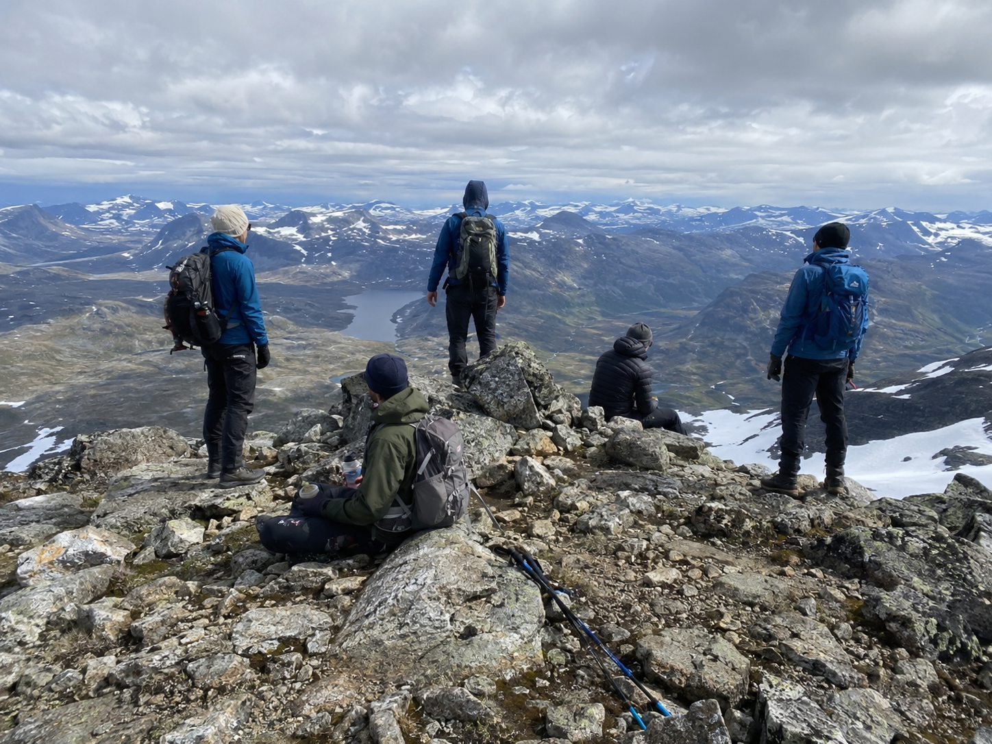 Hikers resting and taking in the view from a rocky mountain summit surrounded by snow-covered peaks and valleys.
